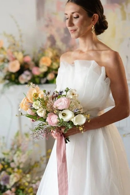 Bride in a white wedding dress holding a bouquet of pastel flowers standing in front of a floral background.