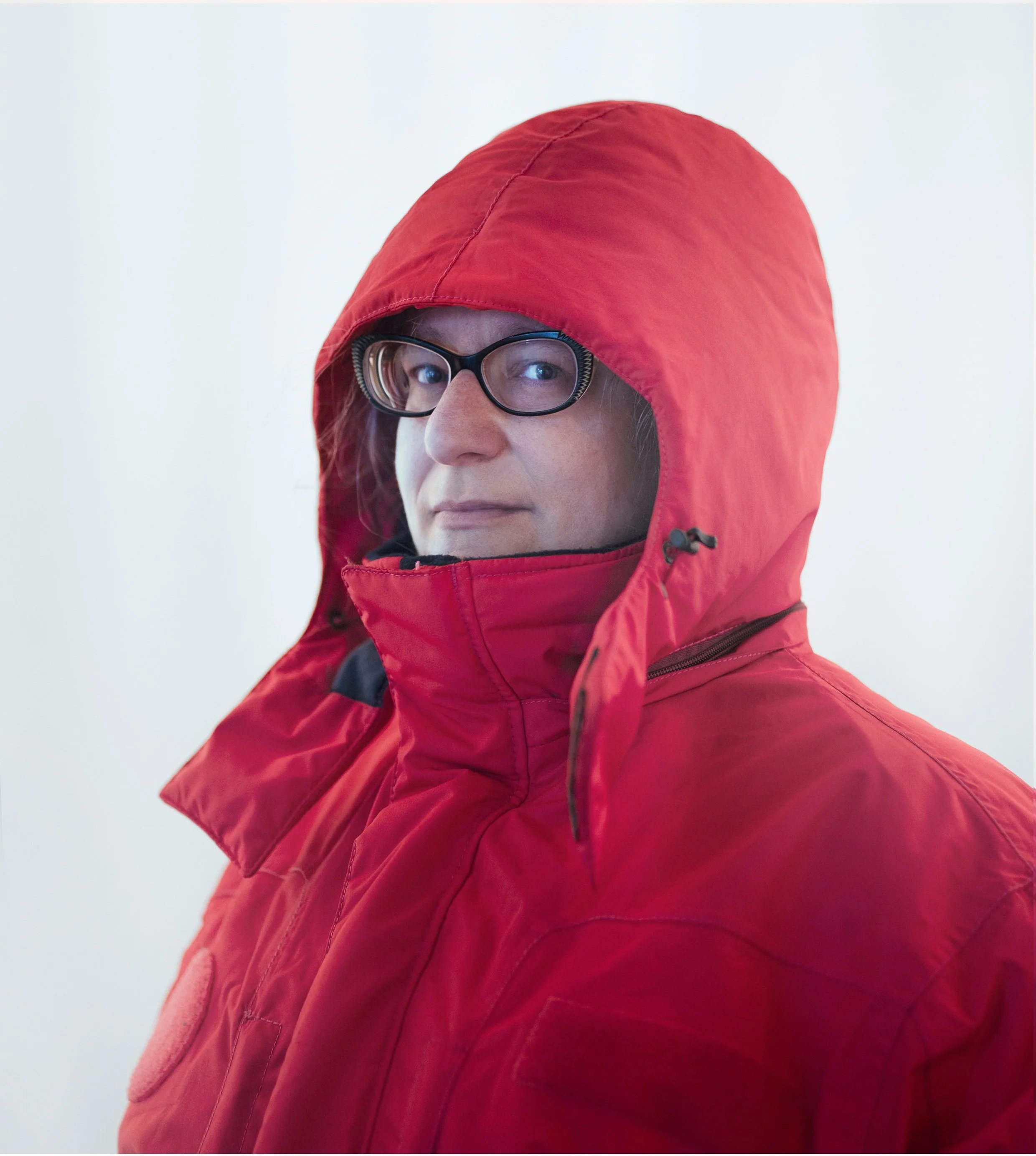 A white woman with dark rimmed glasses. She is wearing a large red parka and is looking sideways toward the camera. She has a slightly cheeky look on her face.