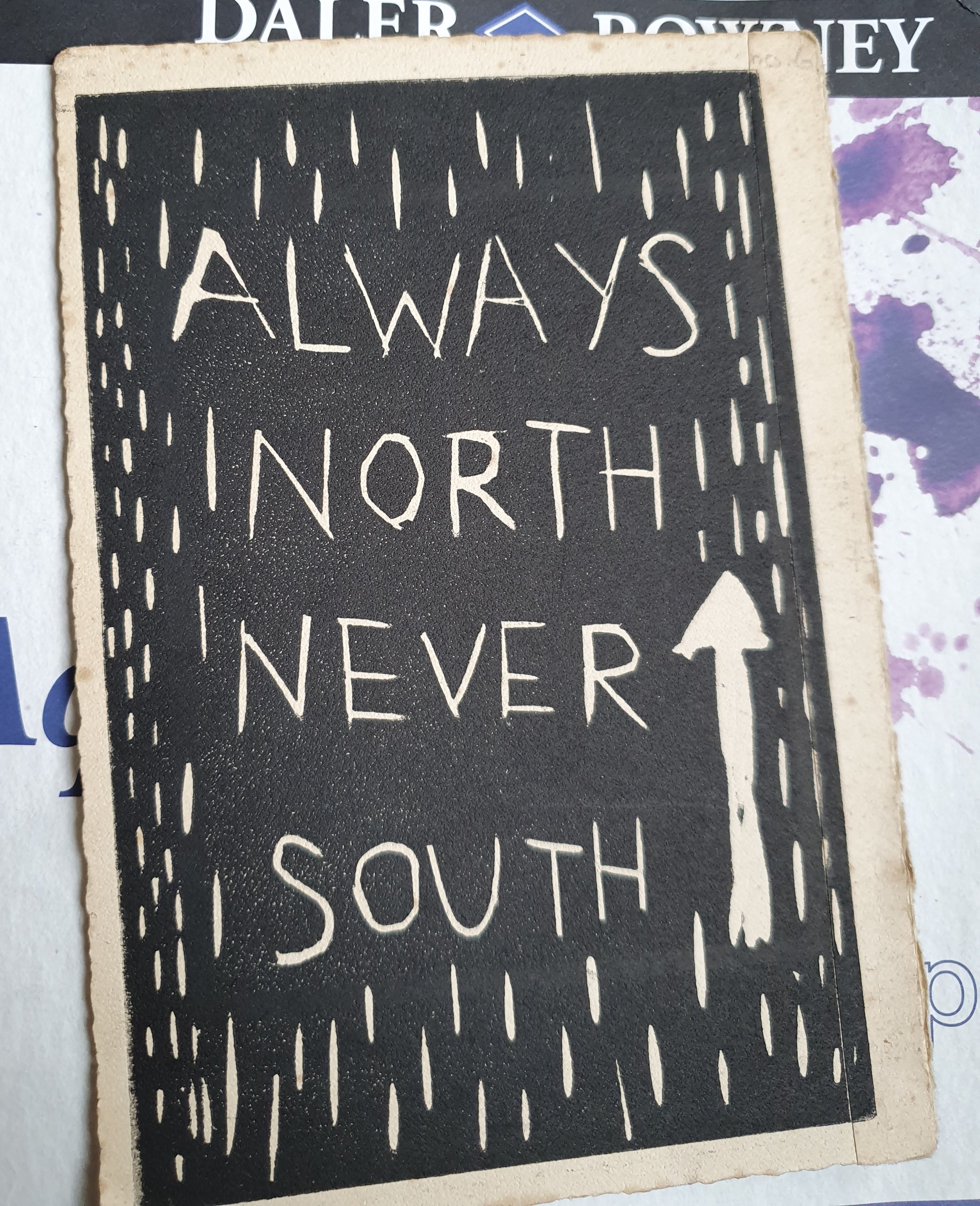A linocut print in black ink on an old book page. The text says 'Always North Never South' there are also marks made running down the print that are reminiscent of rain. There is an arrow pointing upwards.