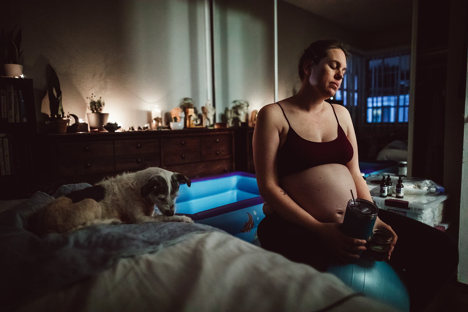 A pregnant woman in a black sports bra sitting on a blue exercise ball in a dimly lit room, holding a drink and a jar, with a small dog lying on the bed nearby.