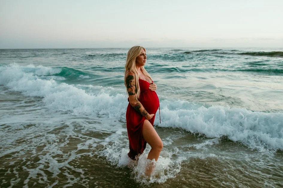 Pregnant woman in a red dress standing in the ocean waves at the beach.
