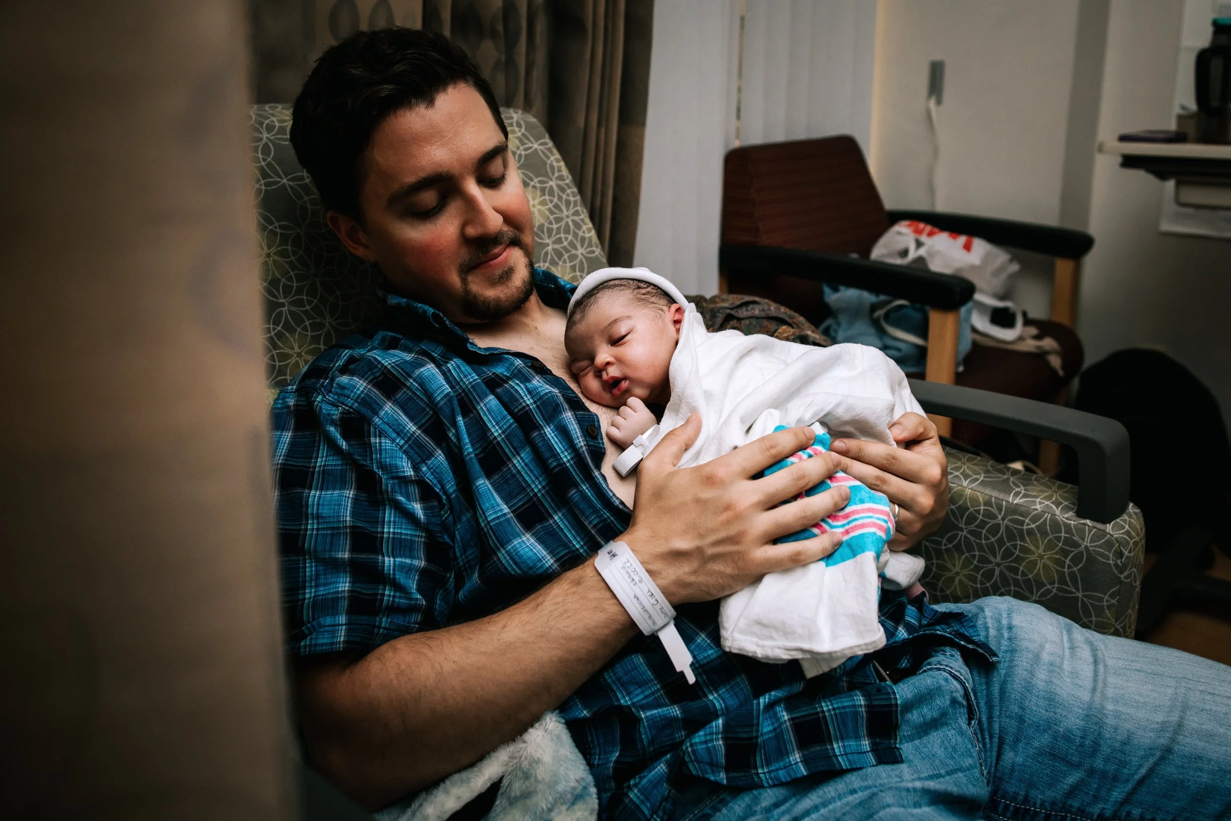 A man in a blue plaid shirt holding a newborn baby wrapped in a white blanket while lying on a hospital bed in a hospital room.