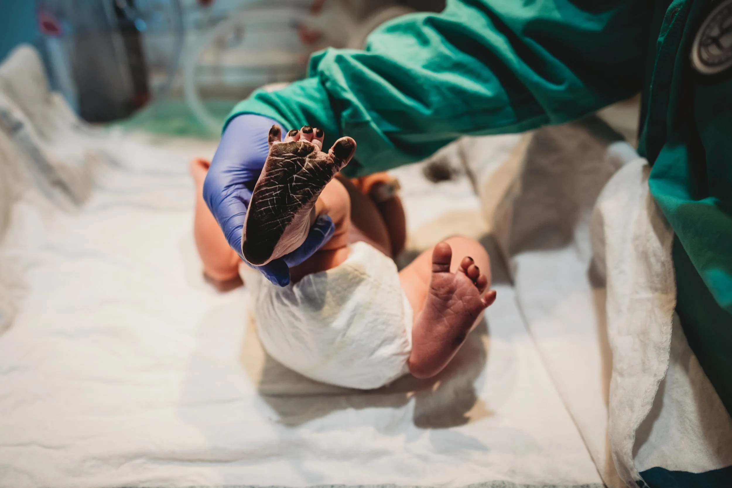 Close-up of a newborn baby lying on a hospital bed with a medical professional holding its tiny foot with gloved hands.