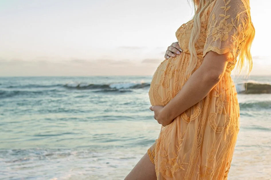 Close-up of pregnant woman in yellow lace dress holding her belly on the beach at sunset.