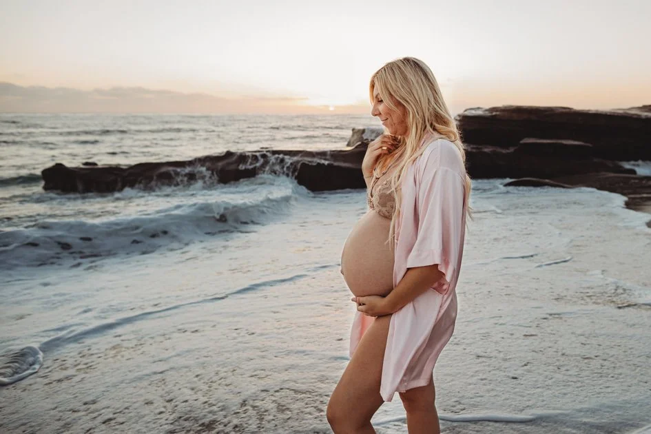 Pregnant woman standing on a beach at sunset, holding her belly and smiling.