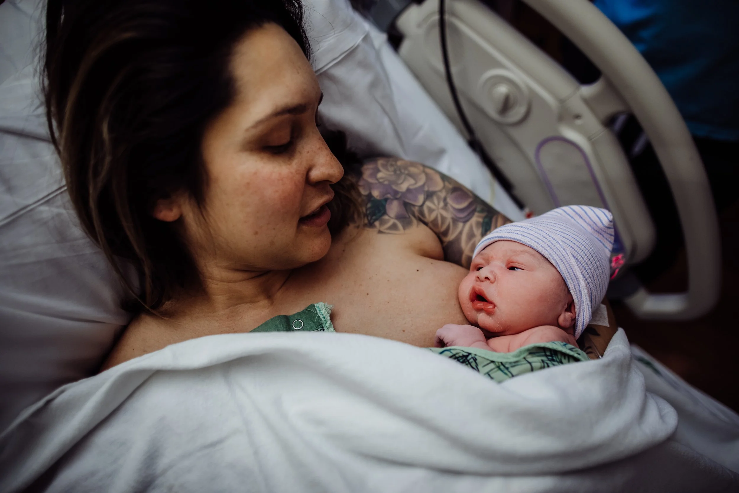 A woman holding a newborn baby in a hospital bed, looking at the baby with affection. The woman has a tattoo on her shoulder and is lying on her side, with the baby resting on her chest, wearing a striped hat.