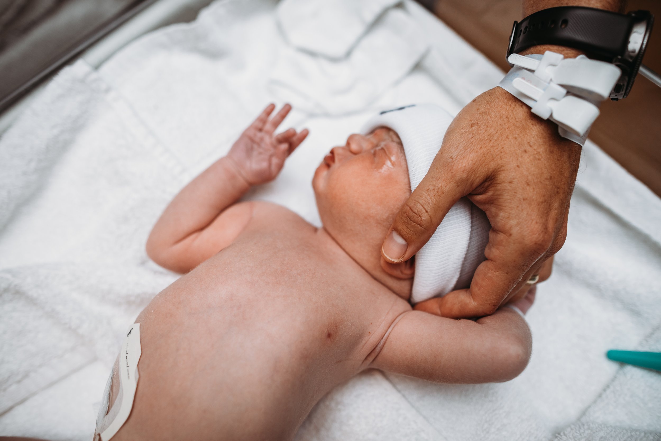 Newborn baby lying on a white towel with an IV line attached, being gently held by an adult hand, likely in a hospital setting.