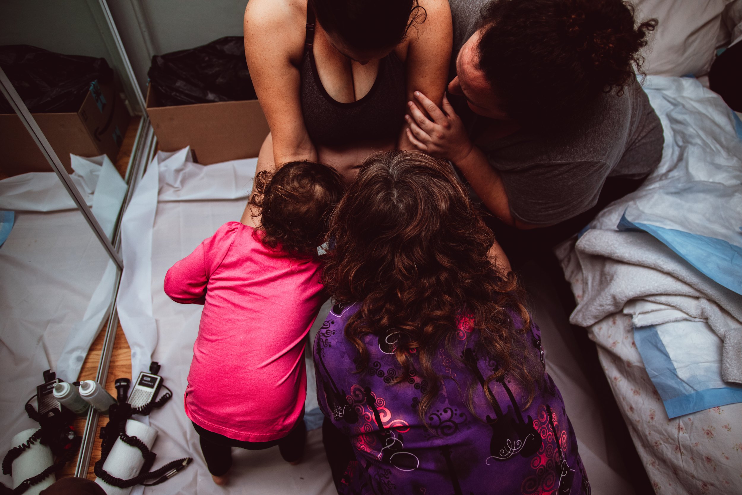 Family of four gathered on a hospital bed, with a woman in labor holding a newborn, and another woman and a young girl leaning in close, all looking at the baby.