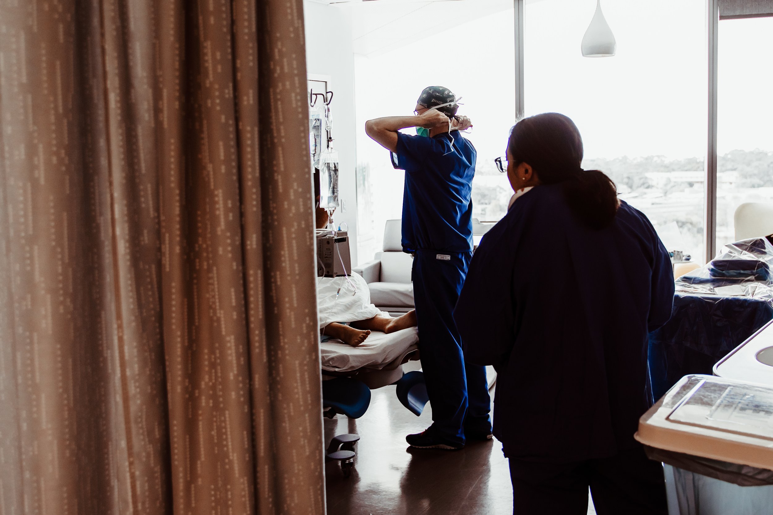 Healthcare workers attending to a patient lying in a hospital bed, with one worker adjusting equipment and another observing, in a hospital room with large windows.