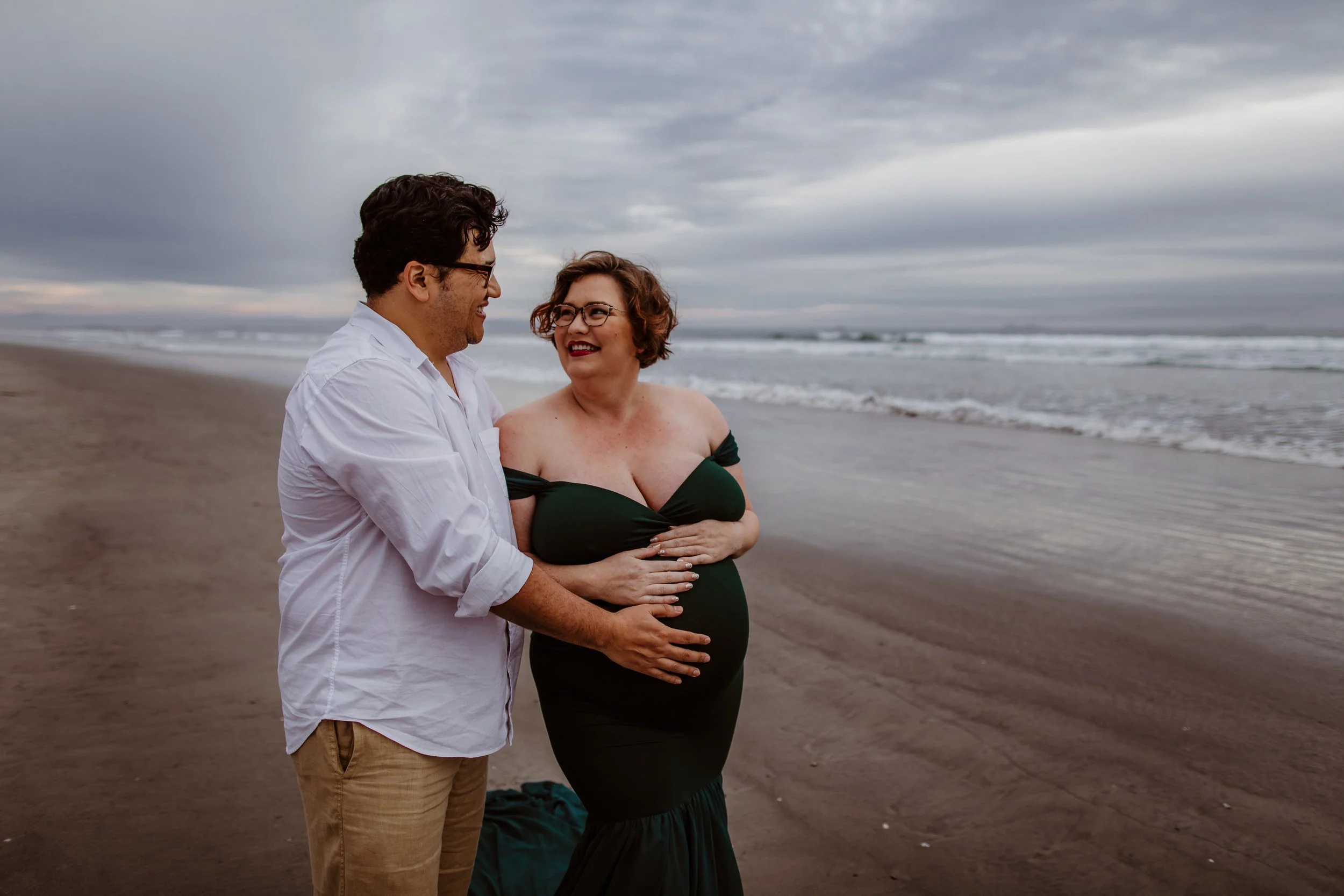 A happy couple stands on the beach, celebrating pregnancy, with cloudy sky and ocean waves in the background.