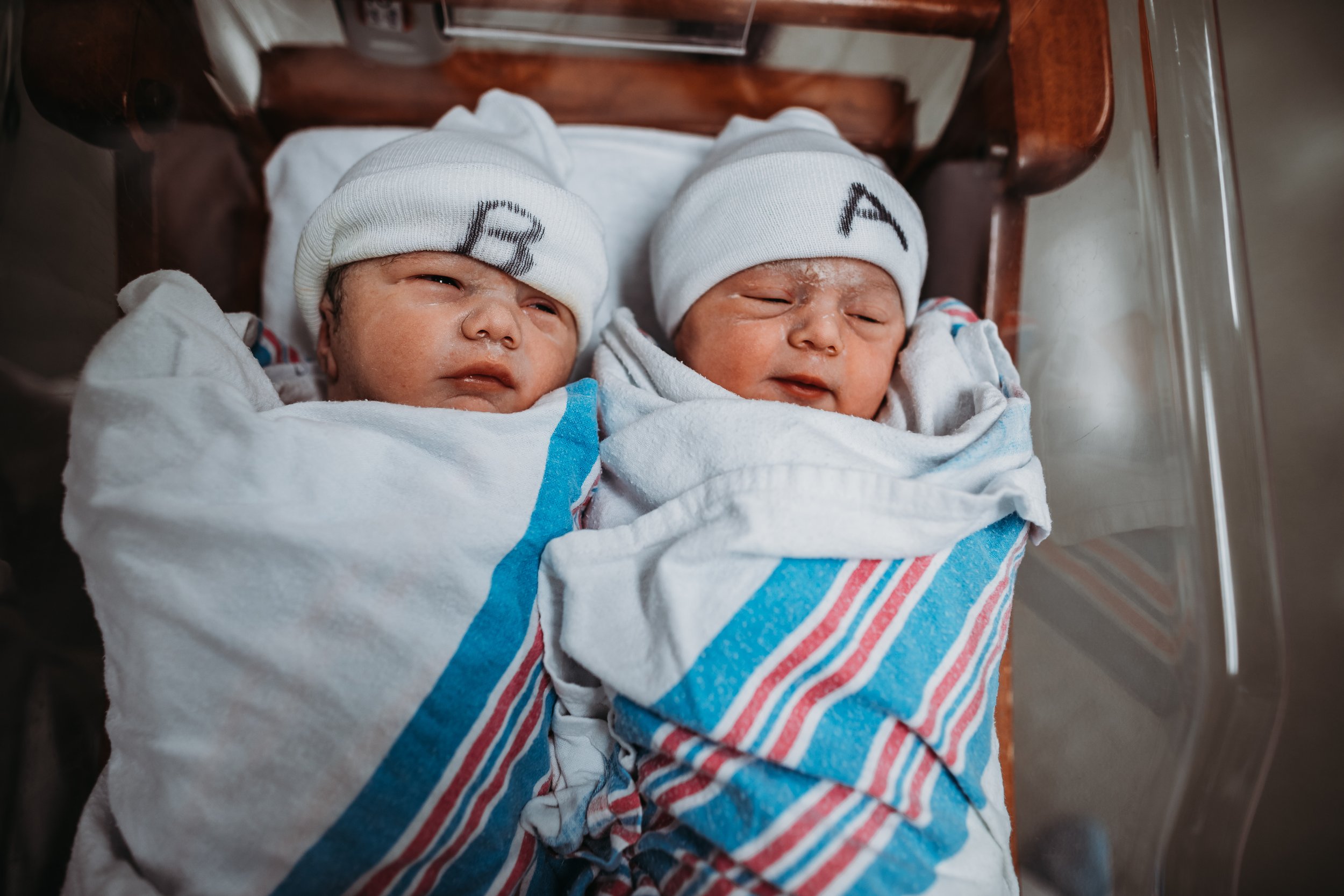 Two newborn babies in a hospital bassinet, swaddled in white blankets with blue and red stripes, wearing white beanies with initials 'A' and 'B' on them.