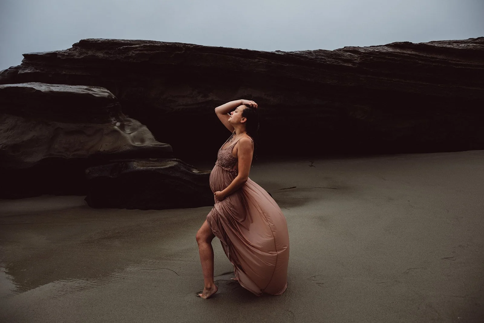 A woman in a pink dress kneels on the sandy beach in front of a large rock formation, with her hand on her pregnant belly and her other hand touching her head.