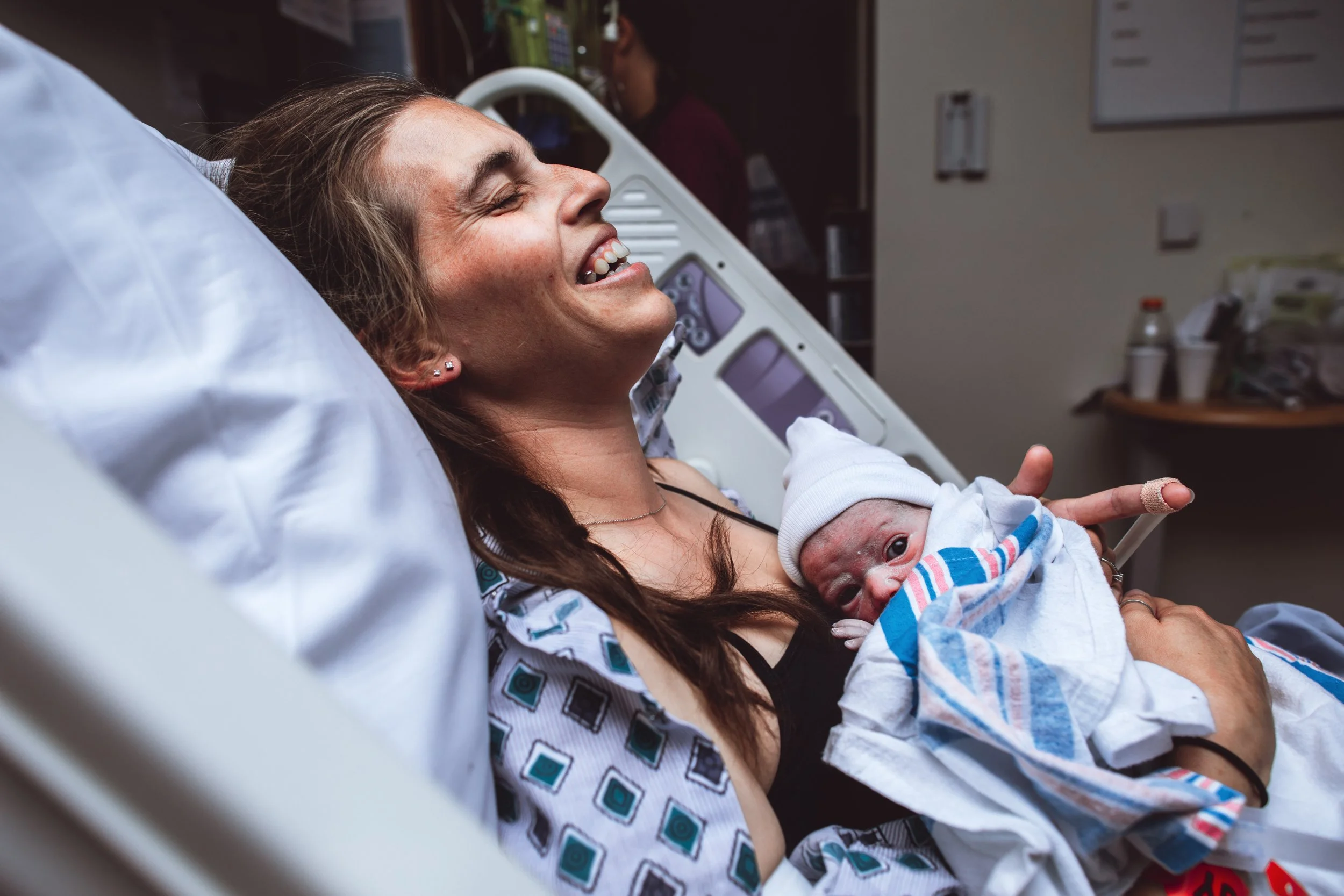 A woman in a hospital bed holding a newborn baby wrapped in a blanket, both smiling.