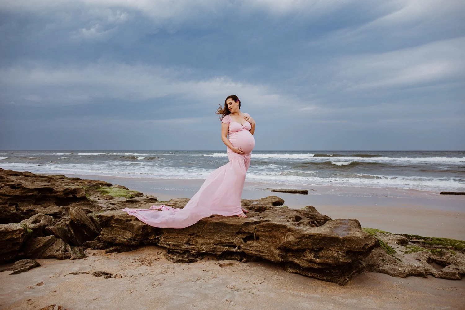 Pregnant woman in a pink dress standing on rocks at the beach with ocean waves and cloudy sky in the background.