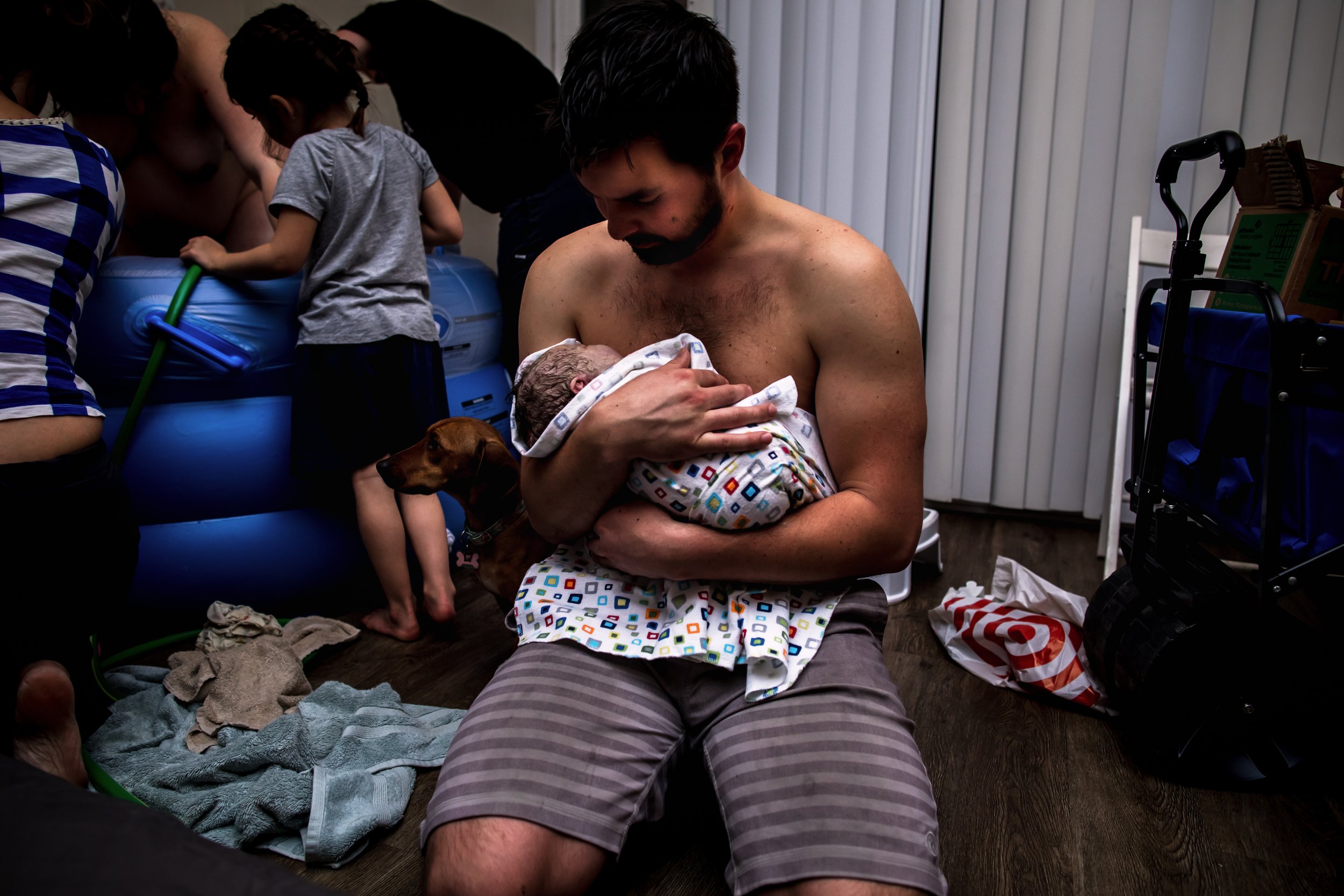 A man holds a newborn baby inside a home, with a dog beside him and children checking on their mother in the birth tub.