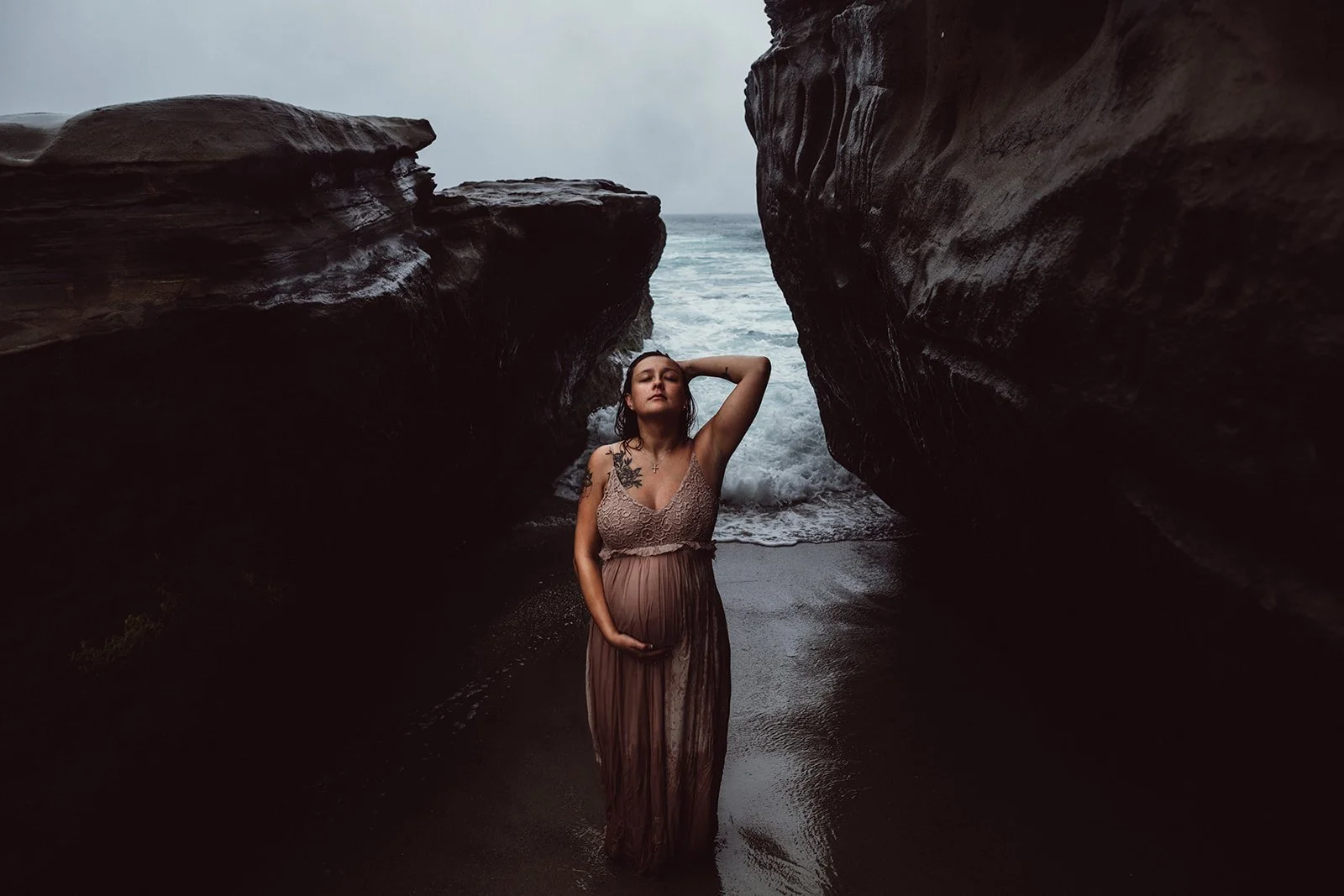A pregnant woman standing between large rocks with the ocean and waves in the background, wearing a sleeveless dress.