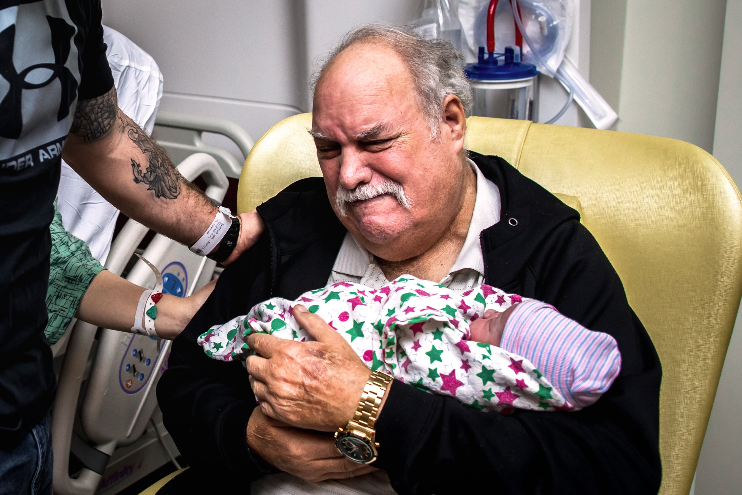 An elderly man with white hair and a mustache, sitting in a hospital chair, holding a newborn baby wrapped in a colorful star-patterned blanket and a striped hat.
