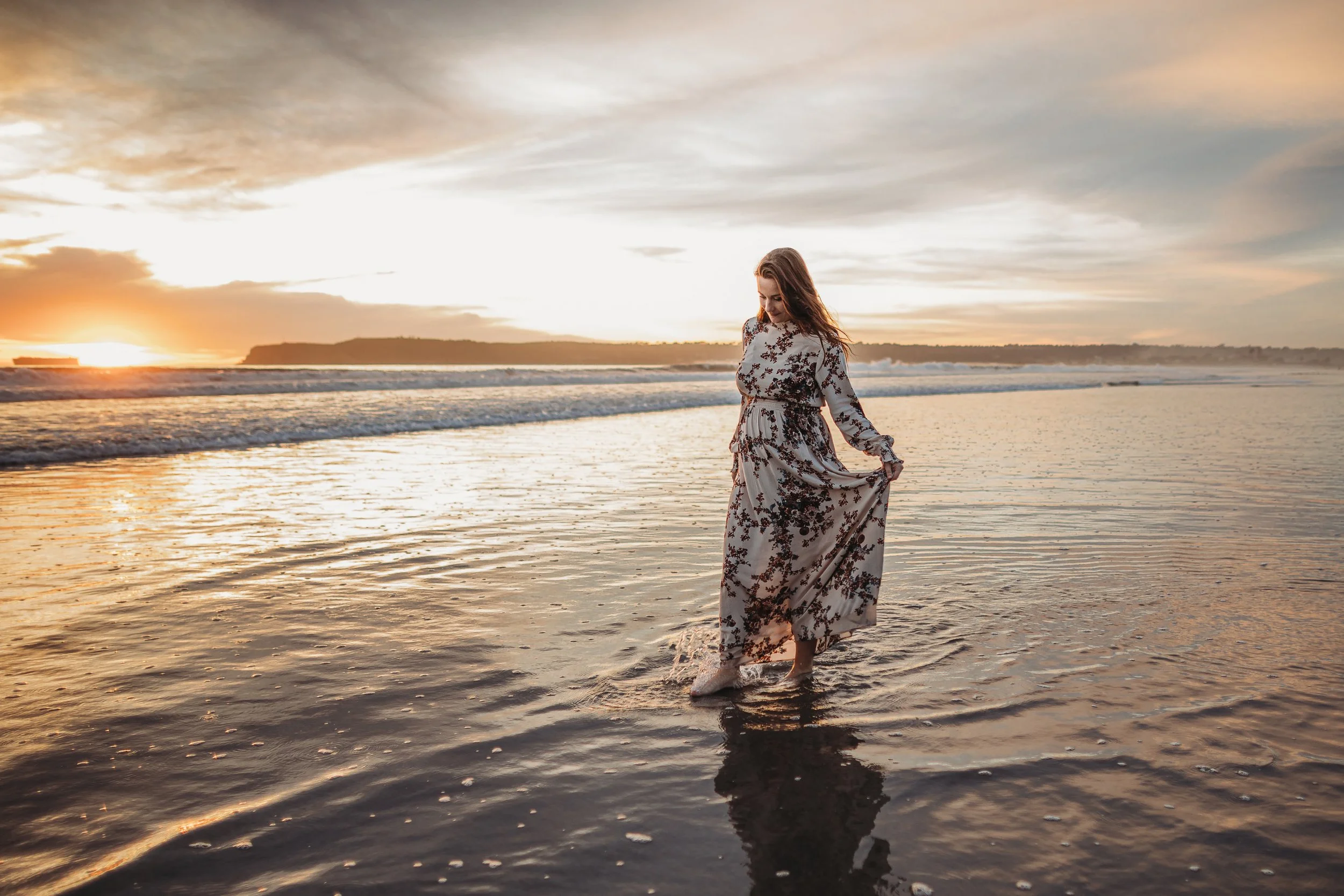 A woman in a floral dress walking in shallow ocean water during sunset