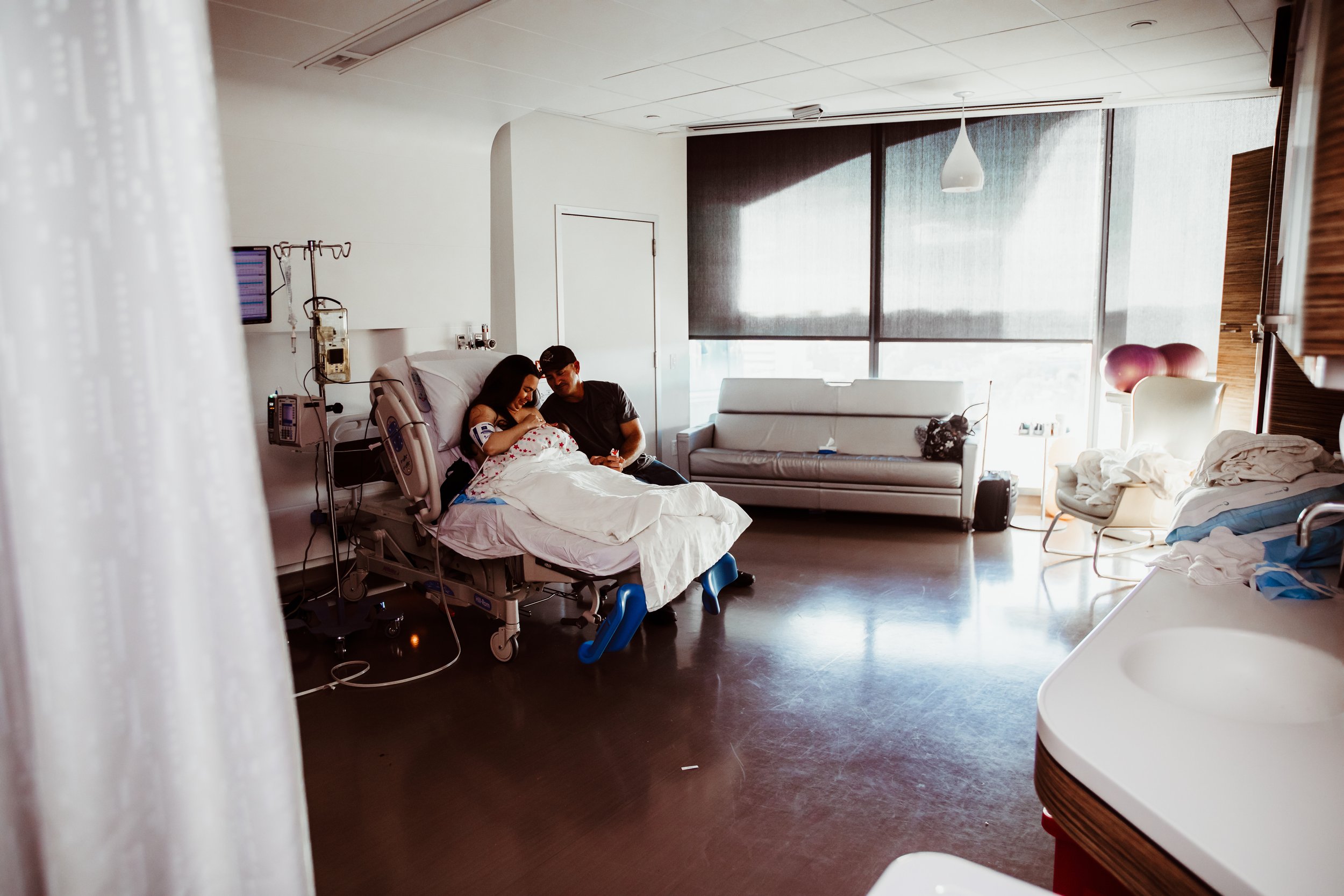 A couple sitting beside a woman in a hospital bed in a hospital room with large windows, a couch, chairs, and medical equipment.
