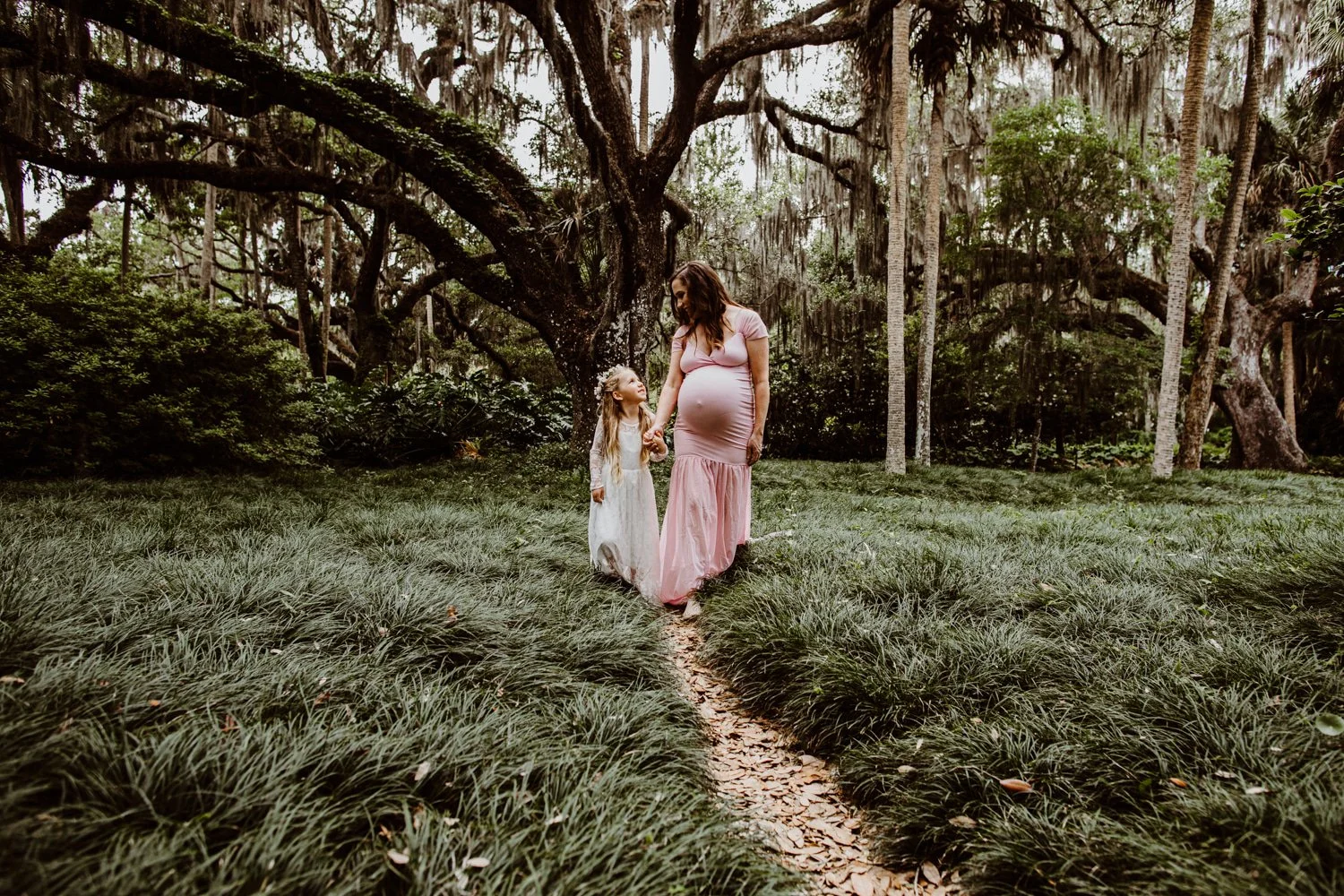 A pregnant woman and a young girl holding hands walking through a forested area with tall trees and green grass.