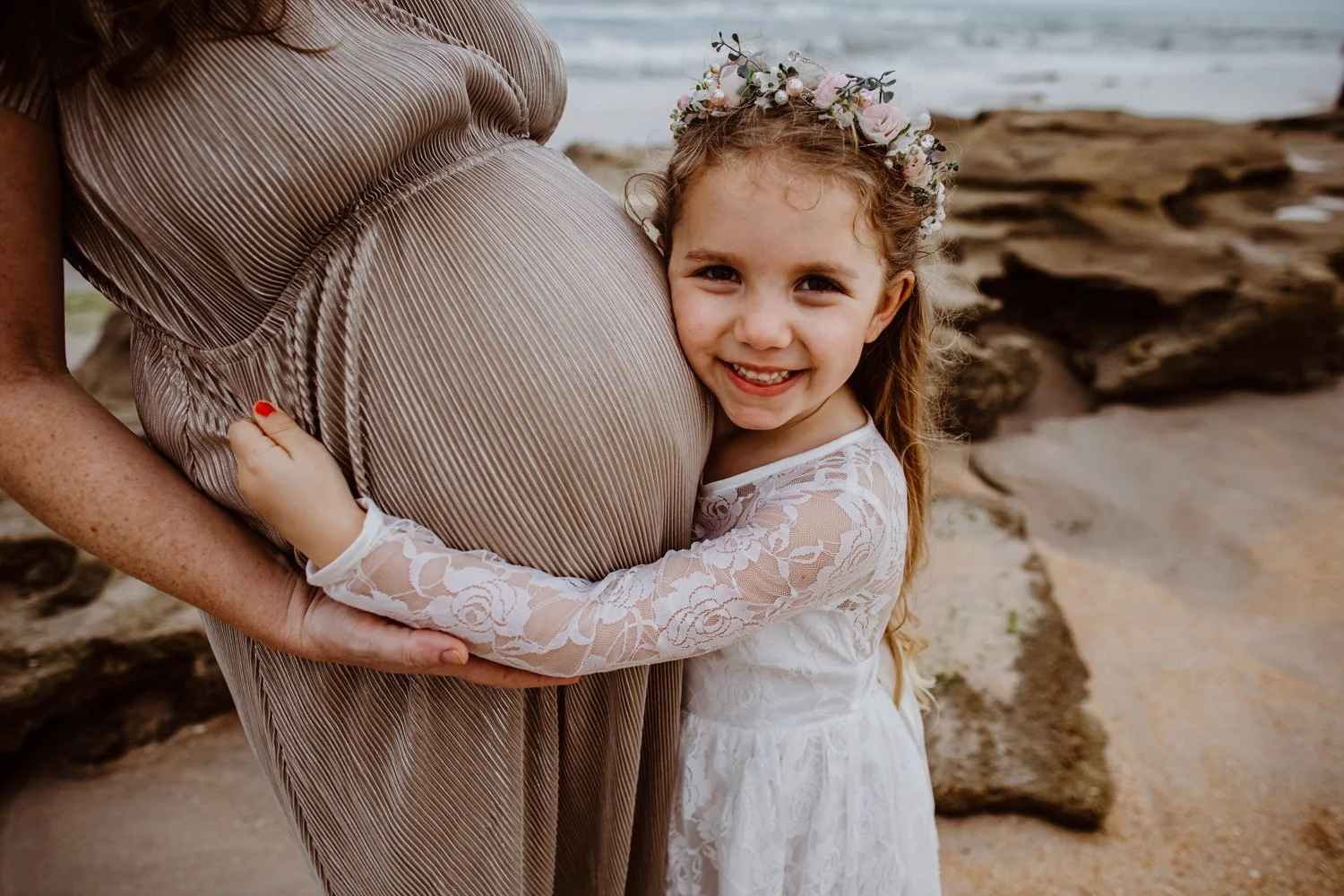 A young girl with a floral headband hugs a pregnant woman on the beach, smiling at the camera.