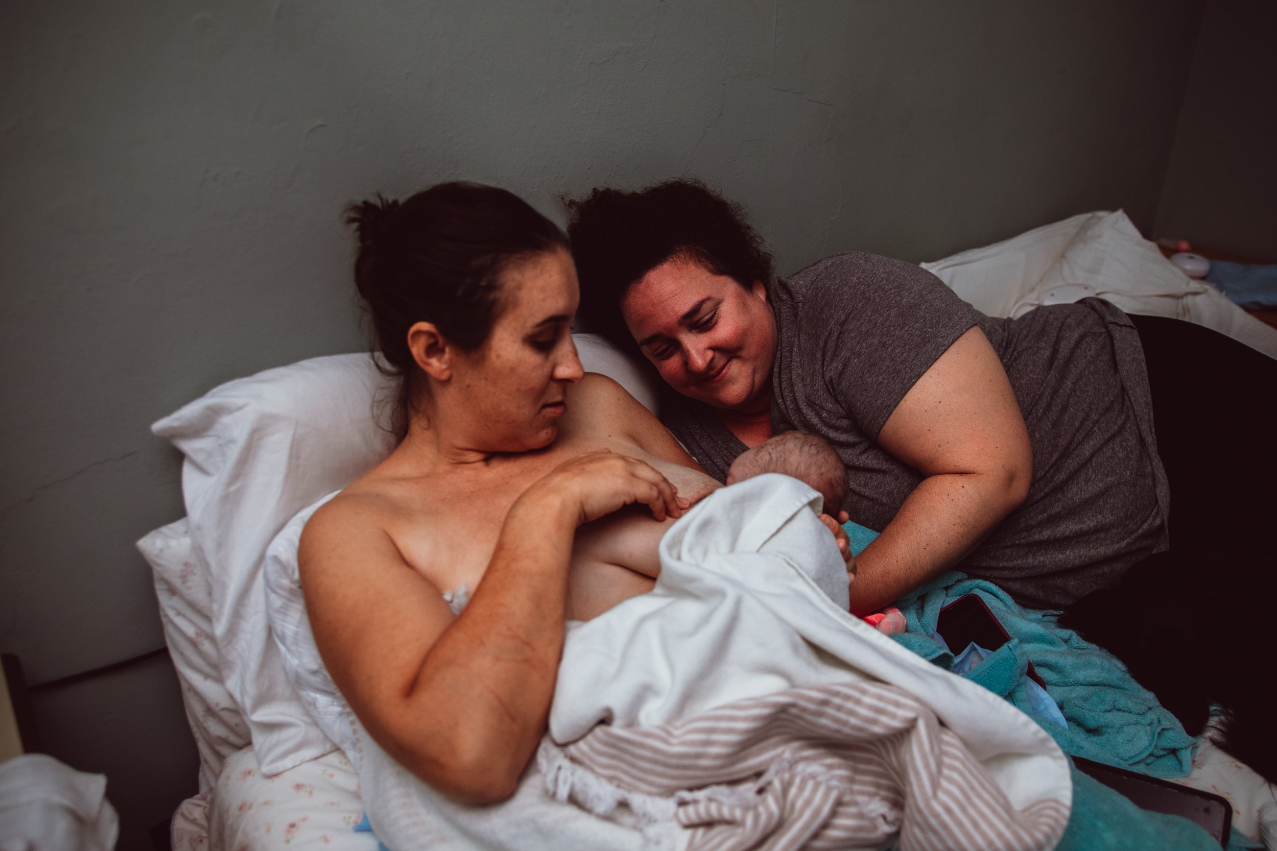 A woman breastfeeding a newborn while another woman leans over, smiling, in a bedroom setting.