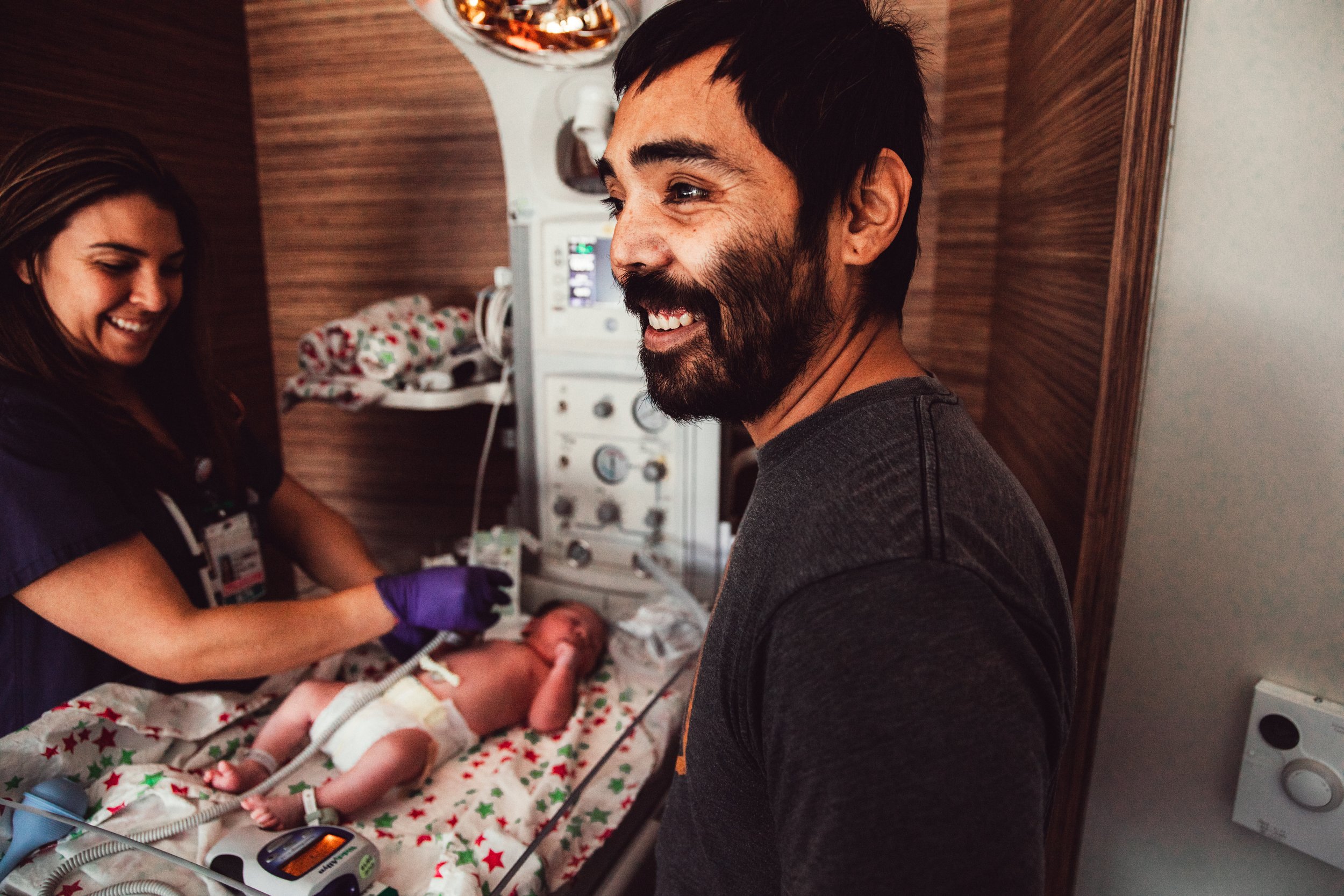 A man smiling as a nurse tends to a newborn in a hospital room.