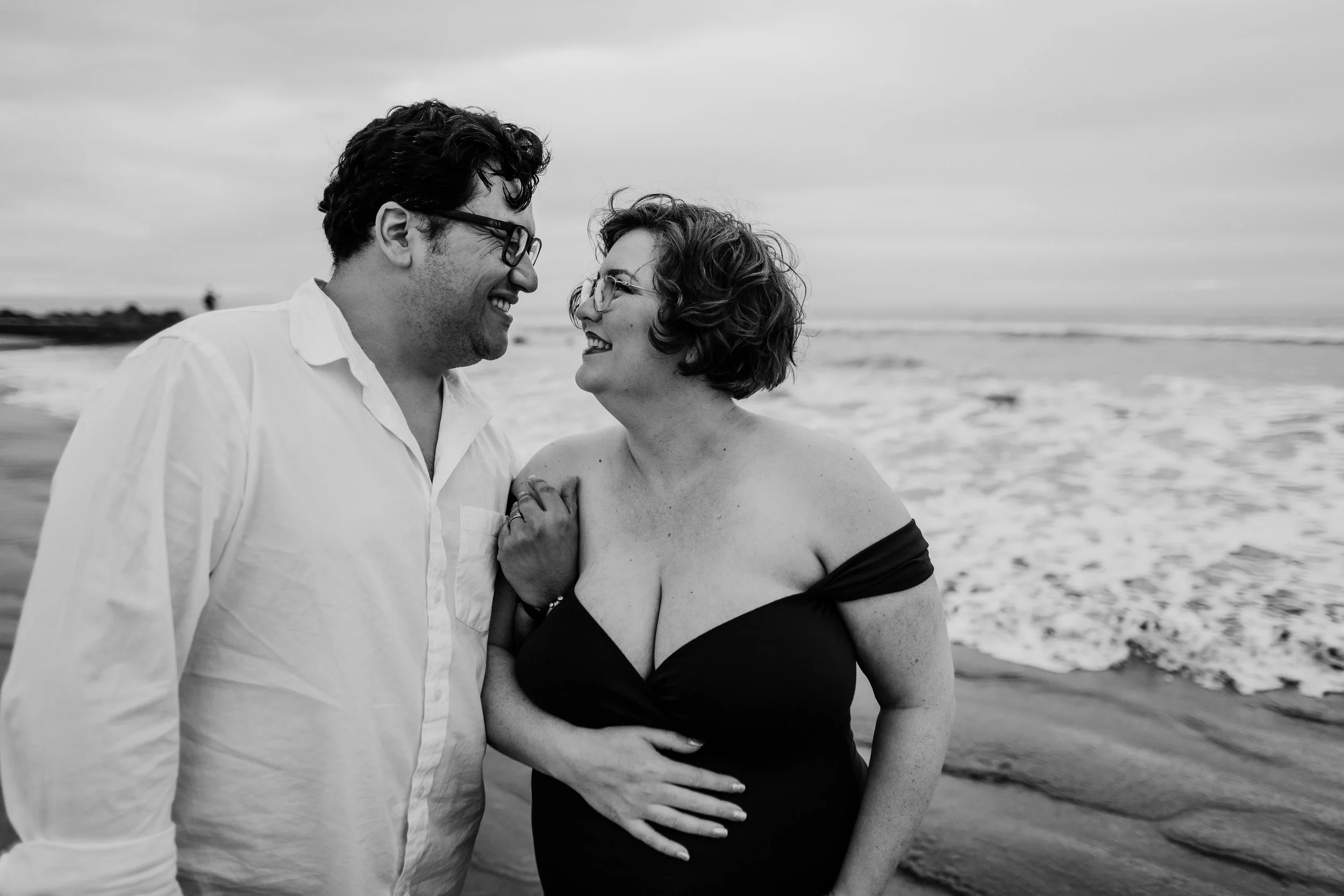 A smiling couple stands on the beach near the water, gazing into each other's eyes, with the ocean and cloudy sky in the background.