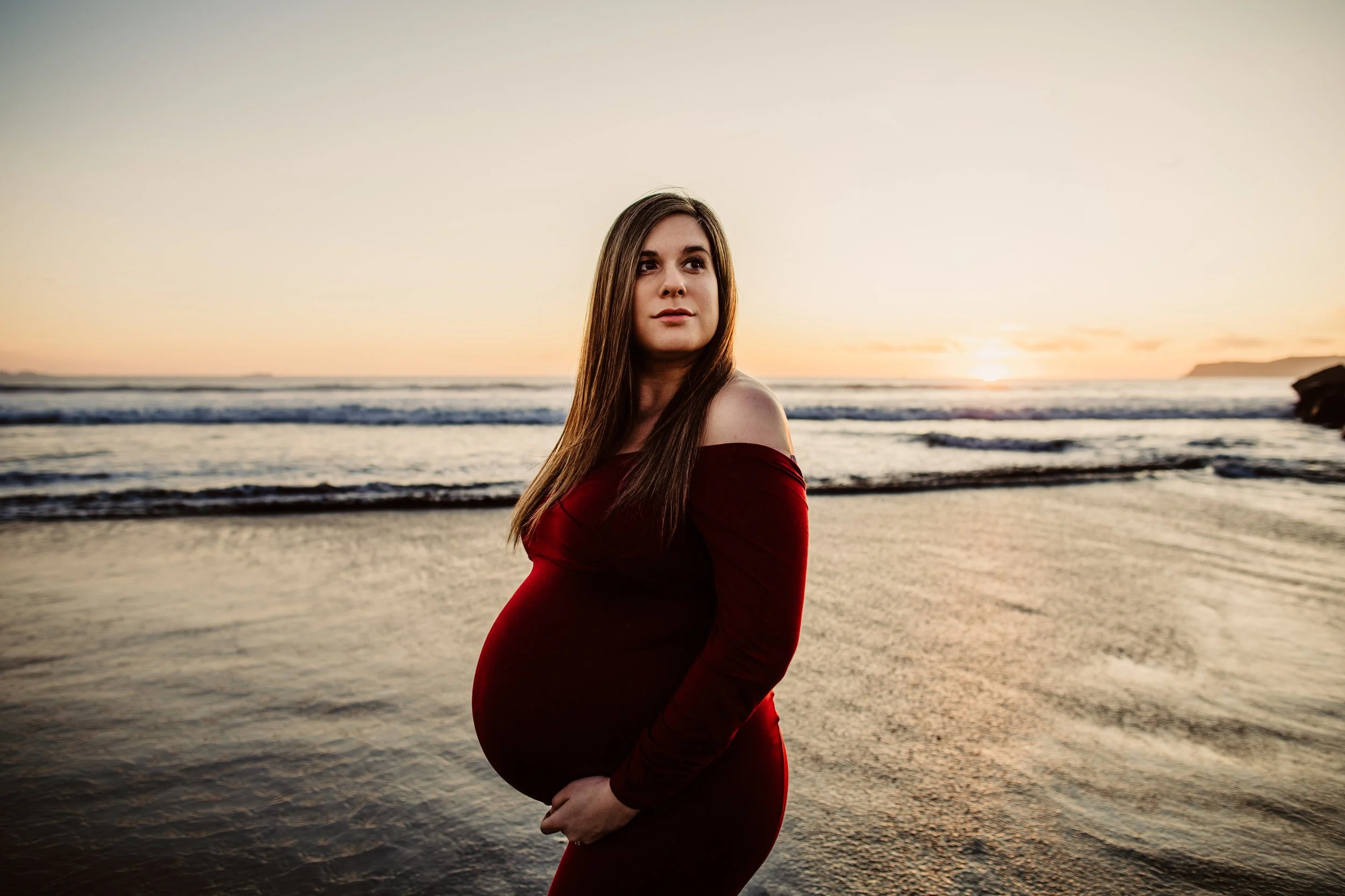 Pregnant woman in a red dress standing on a beach at sunset.