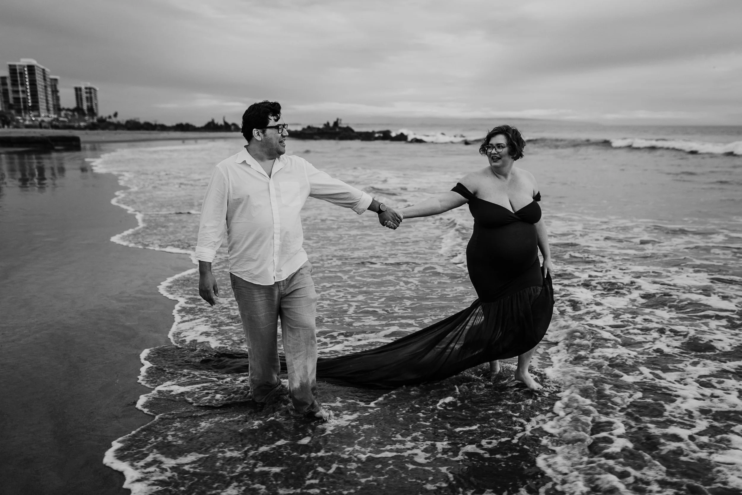A pregnant woman in a dark gown and a man in casual clothing walking hand-in-hand on the beach in black and white.