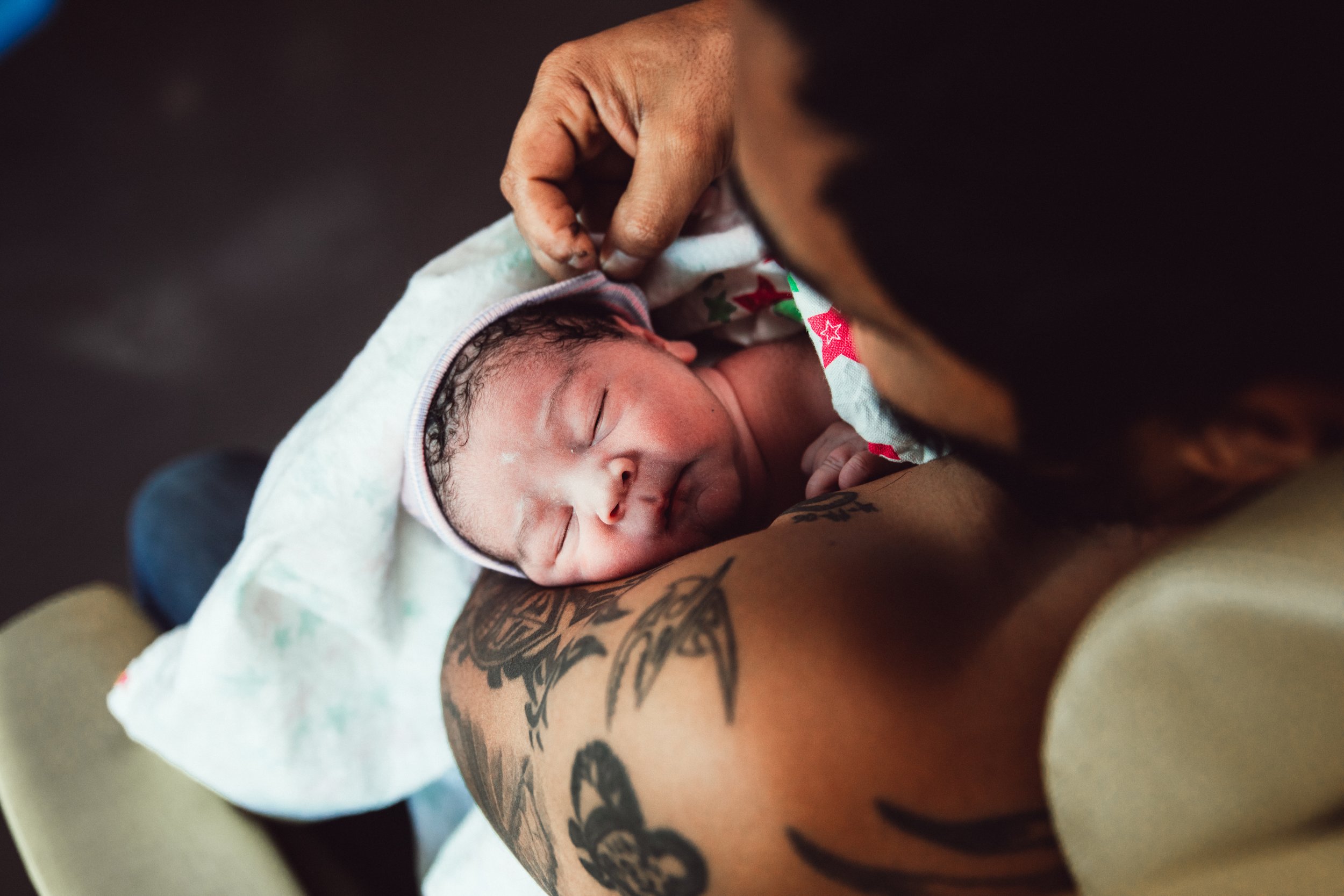 Close-up of a newborn baby lying on a person's chest, covered with a blanket, with the person holding the baby's head gently, showcasing tattoos on the person's arm.