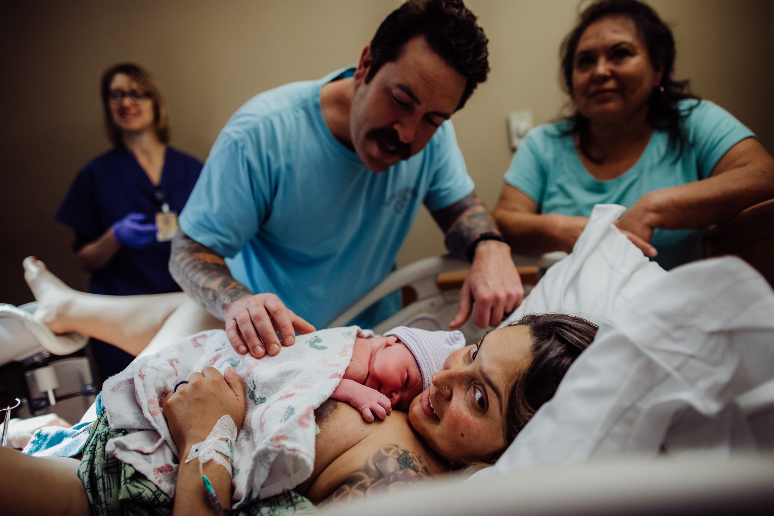 A woman in a hospital bed after childbirth, holding her newborn baby, with a man and a woman beside her, and a nurse in the background.