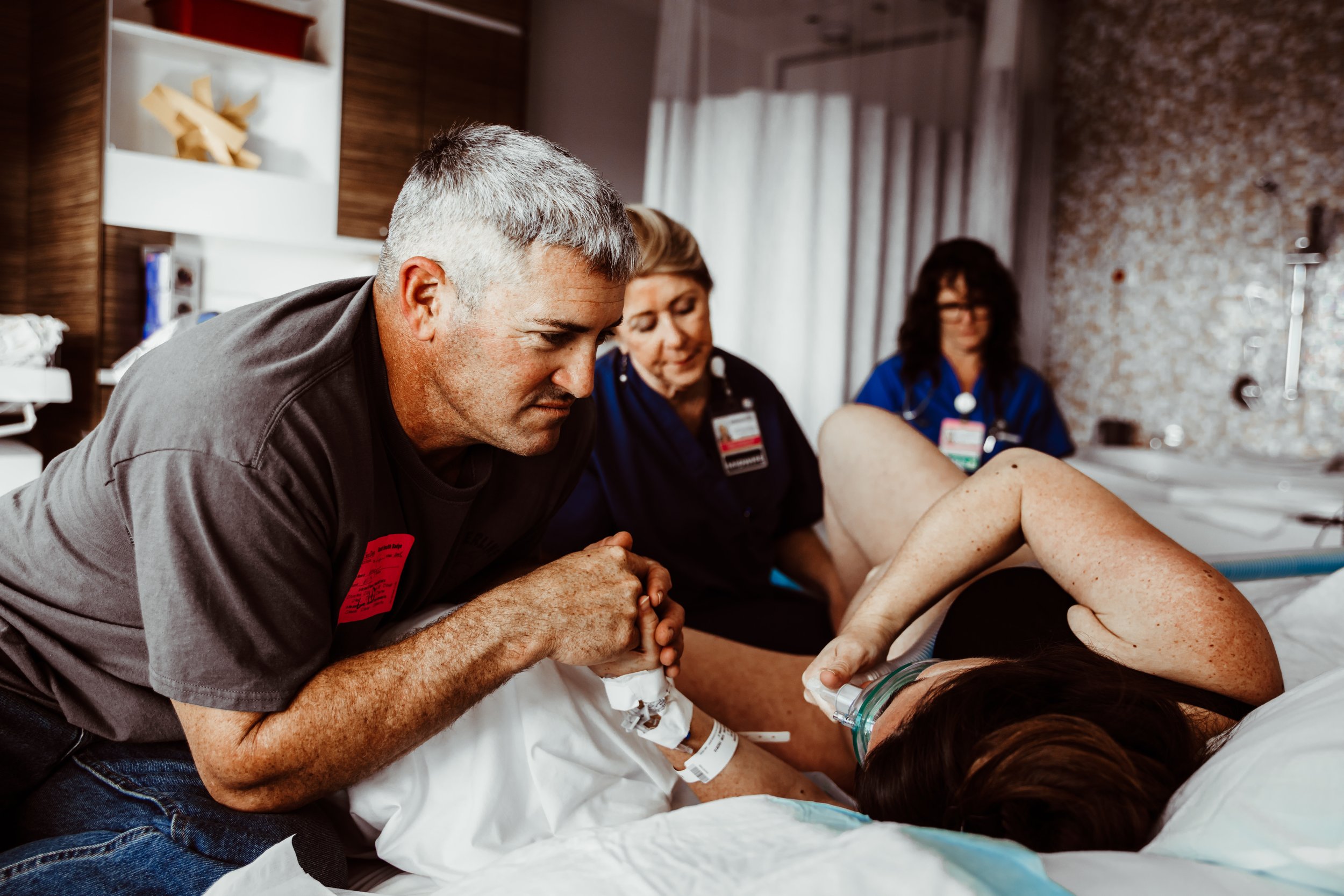 A man in a gray shirt holding the hand of a woman lying in a hospital bed, surrounded by two nurses or healthcare professionals. The woman is giving birth.