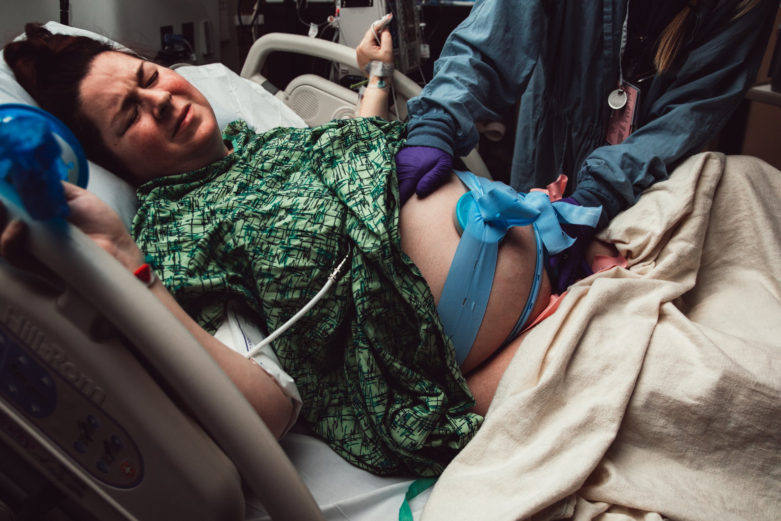 A woman lying in a hospital bed with a distressed expression, connected to medical devices with a healthcare worker holding her hand during childbirth.