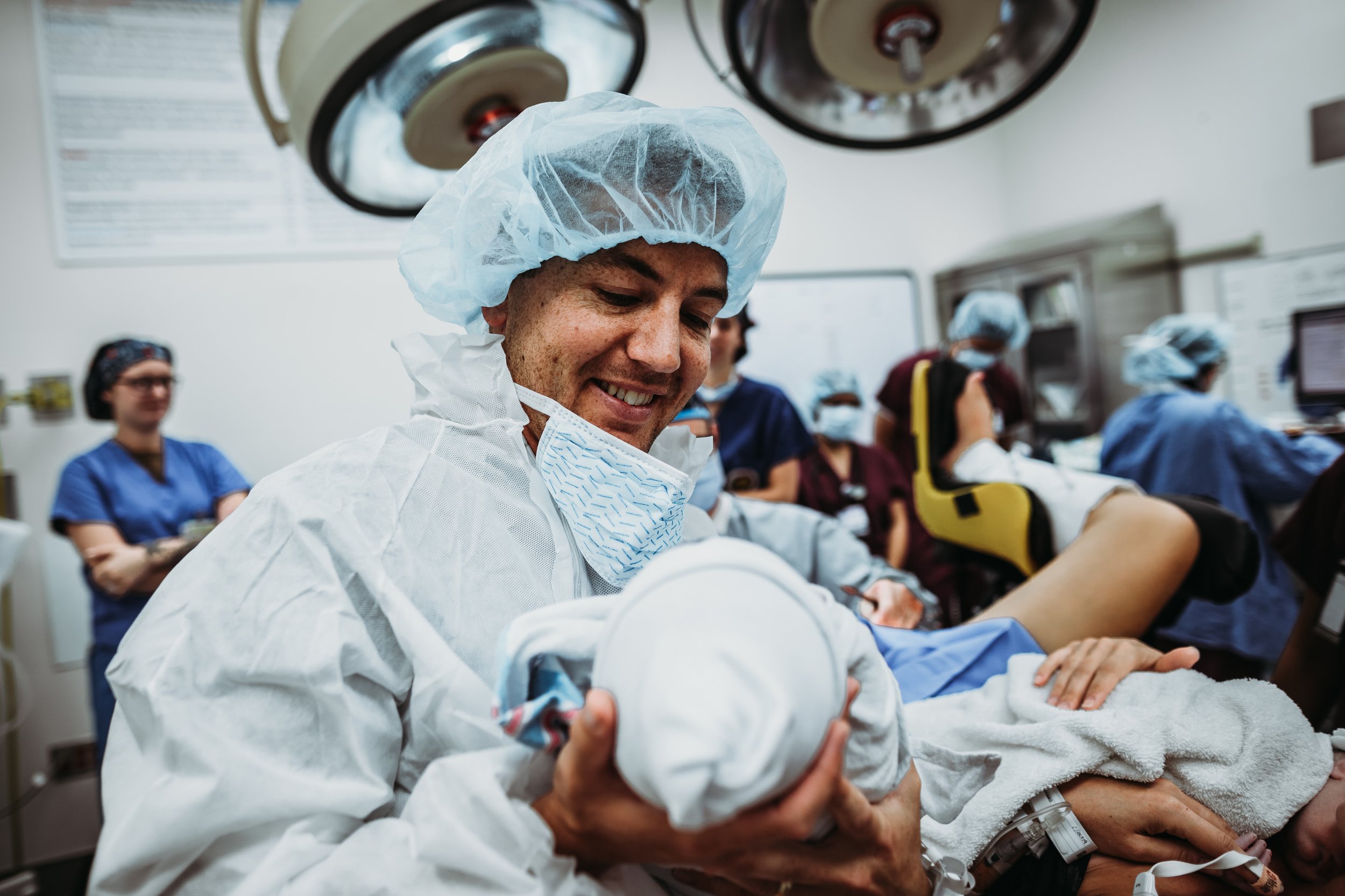 A smiling healthcare worker in surgical attire holding a newborn baby in a hospital delivery room with other medical staff in the background.