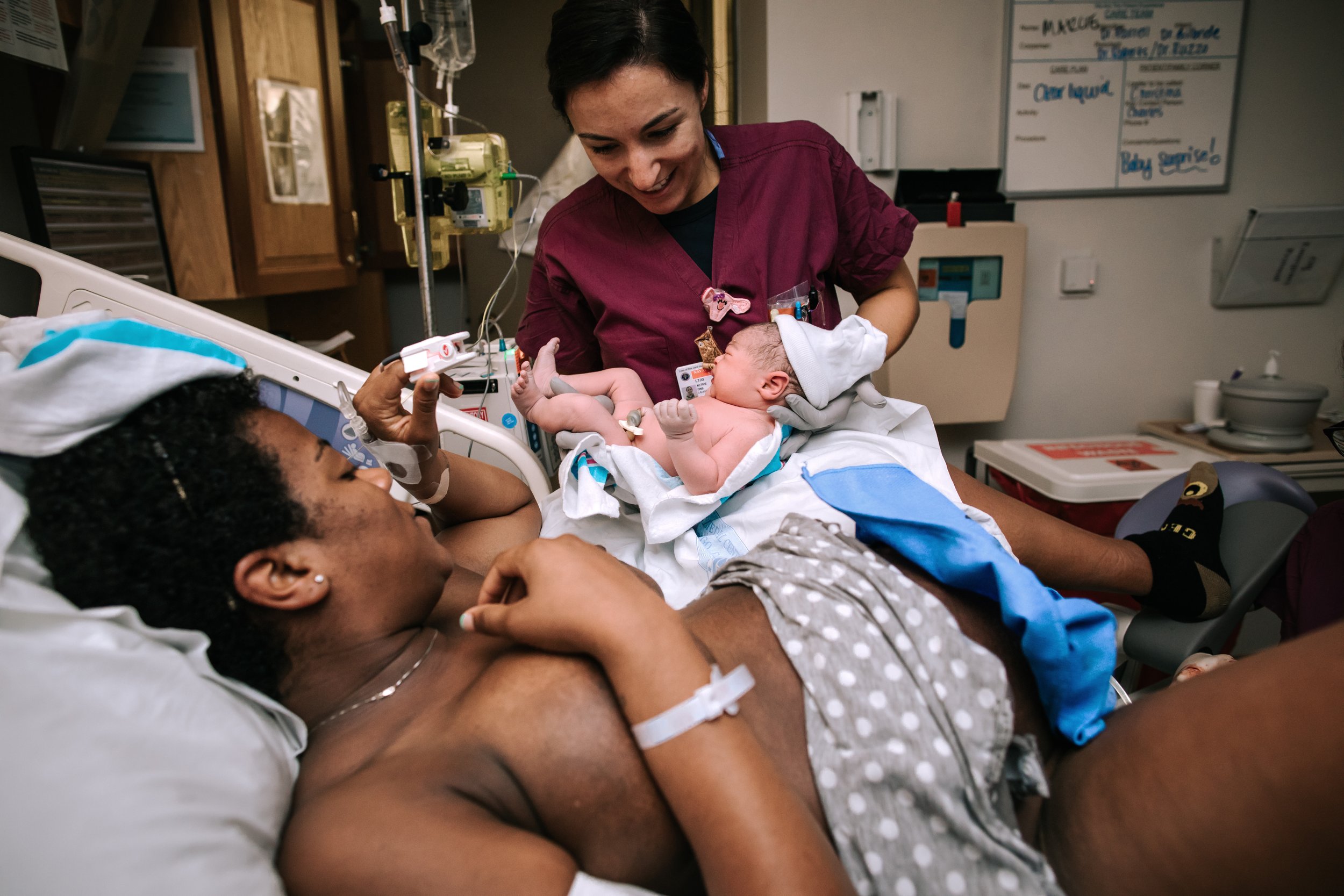 A woman lying in a hospital bed with a newborn baby on her chest, a nurse standing nearby holding the baby, inside a hospital room.