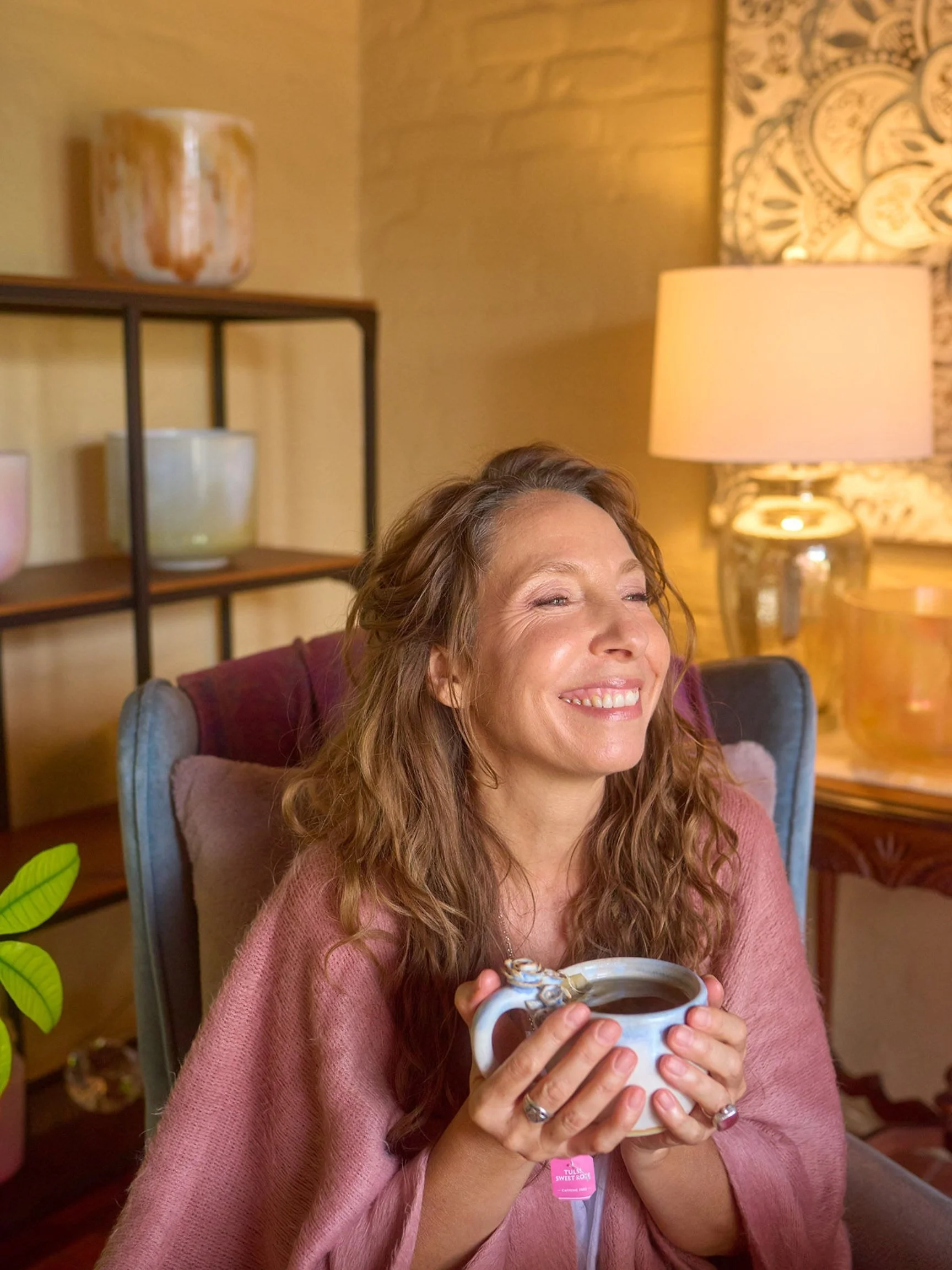 Photo of Amala Rose, SEP, Somatic therapy, Intuitive Channel and Healing Arts, energy and sound healing practitioner in a cozy office with a mug of tea and wearing a soft pink wrap, with crystal singing bowls in the background