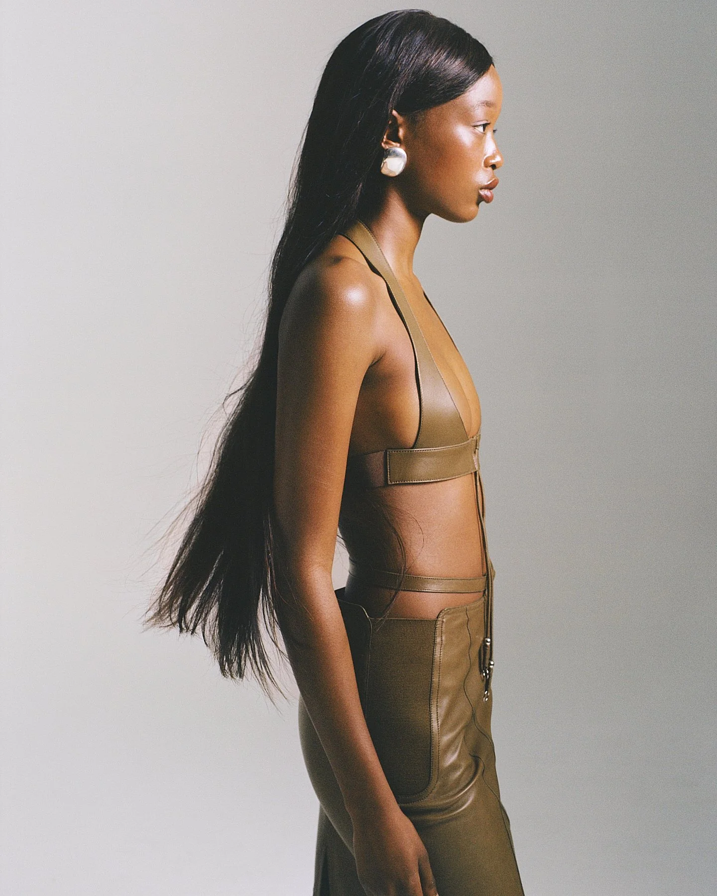 A woman with long dark hair styled straight, wearing a brown leather outfit with structured design and zippers, standing in profile against a plain gray background.