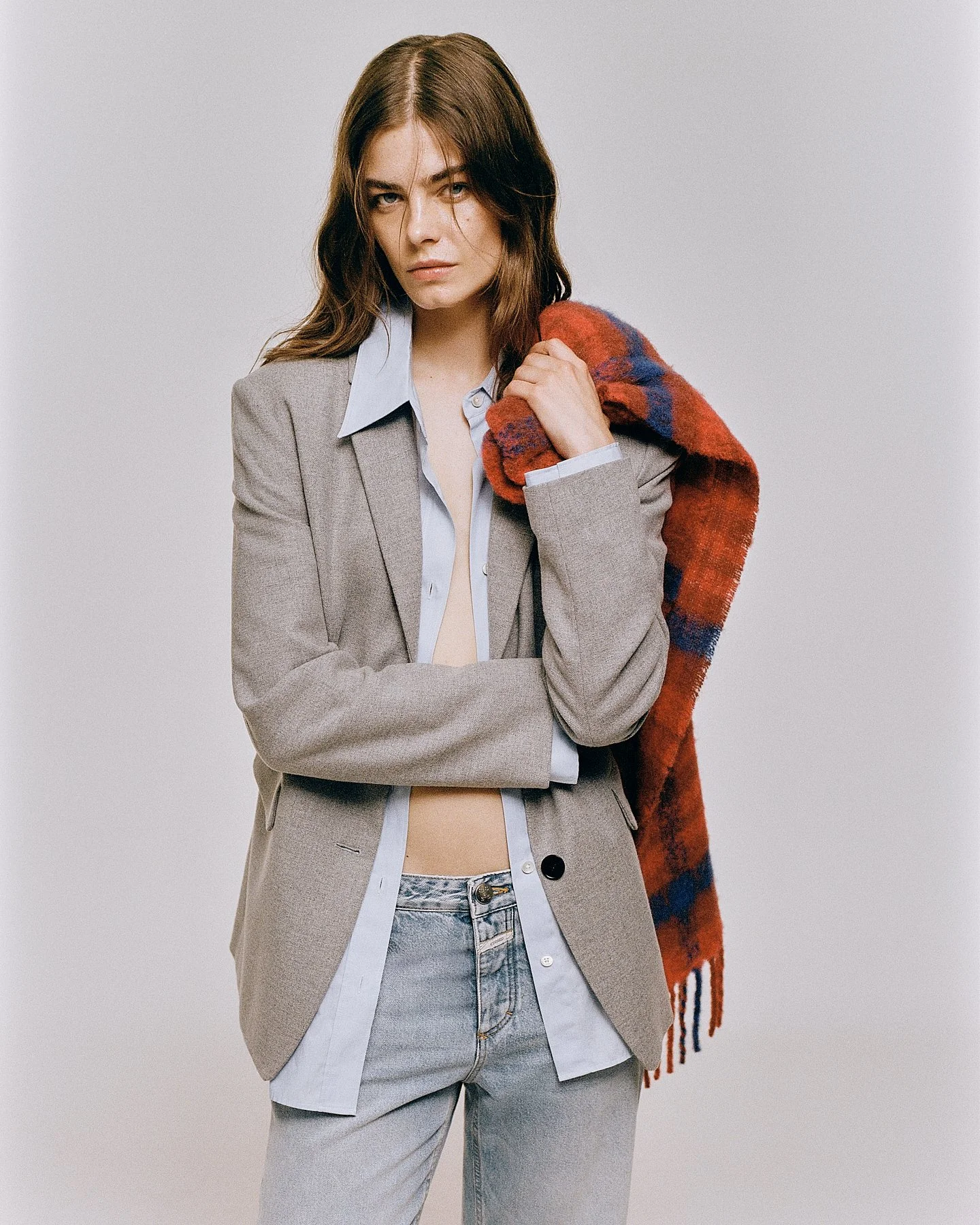 A young woman with brown hair in a blazer, shirt, and jeans, holding a red and blue plaid scarf over her shoulder, against a plain gray background.