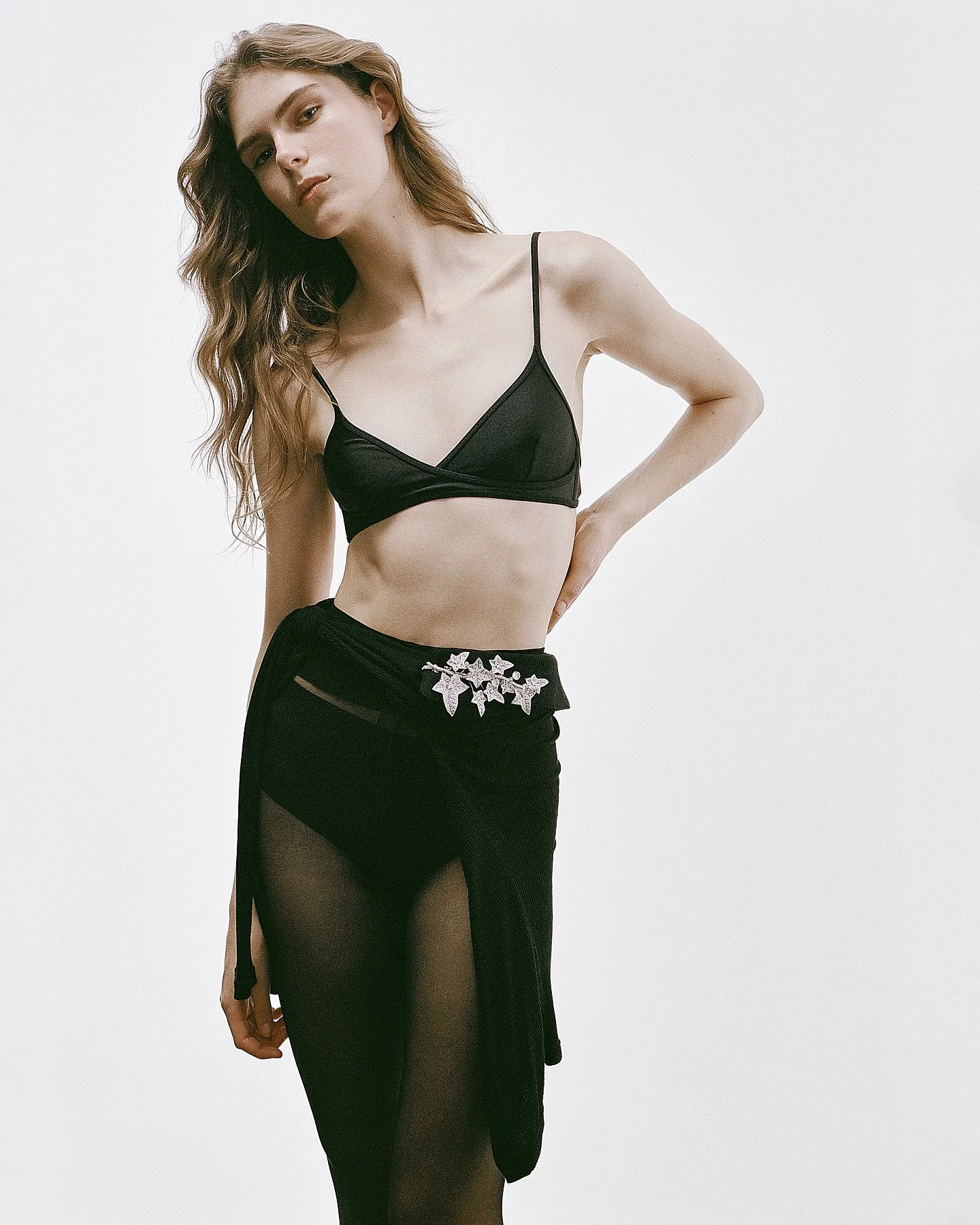 A young woman with long, wavy hair wearing a black cropped bralette top and a black sheer skirt with floral embroidery and side slits, standing against a plain, light-colored background.