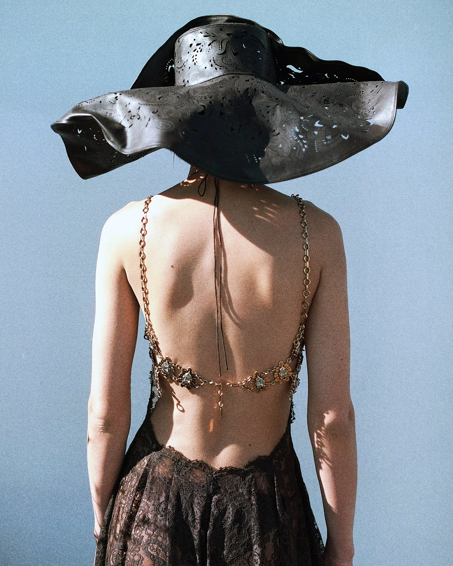 A person wearing a large, black, floppy hat with cut-out decorative details, a gold chain body harness, and black lace skirt, standing against a blue sky.
