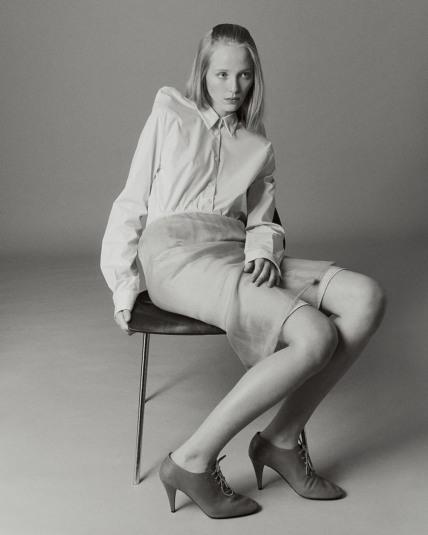 Black and white photo of a woman sitting on a chair. She wears a white shirt, a skirt, and high-heeled ankle boots. Her hair is shoulder-length and styled straight, and she looks thoughtfully at the camera.