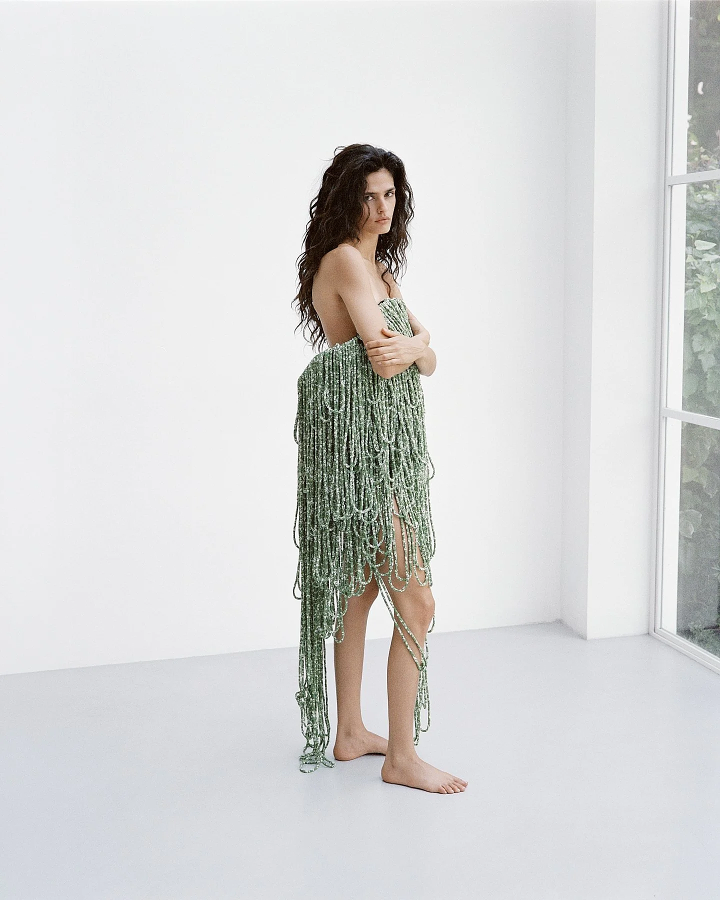 A woman standing barefoot in a white room with large windows, wearing a green dress made of a beaded or fringed material that covers her chest and extends to her calves, with her arms crossed and looking at the camera.