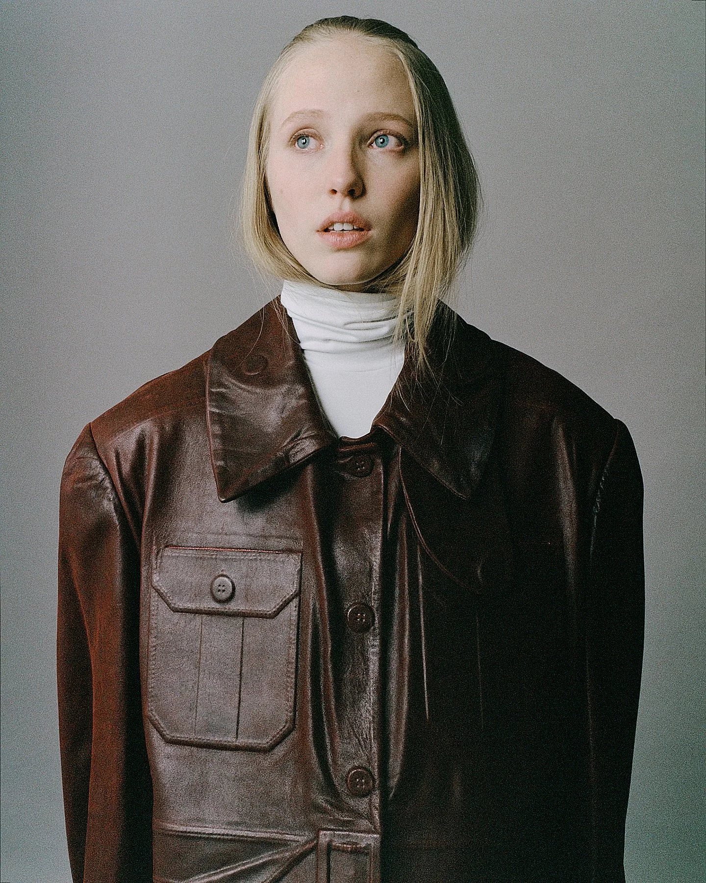 A young woman with blonde hair and blue eyes wearing a white turtleneck and a brown leather jacket, looking slightly to the side against a plain background.