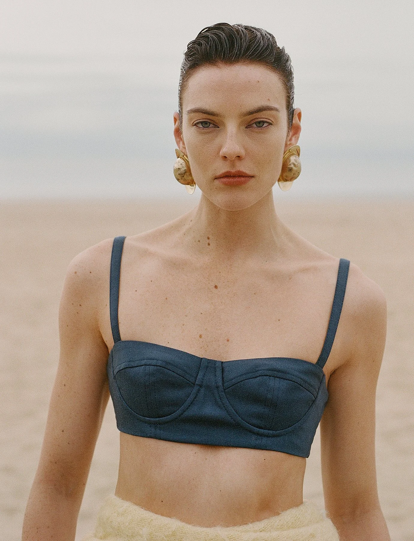 A woman with dark, slicked back hair, wearing large gold earrings, a navy blue strapless top, and a light-colored skirt or towel, standing outdoors against a neutral background.