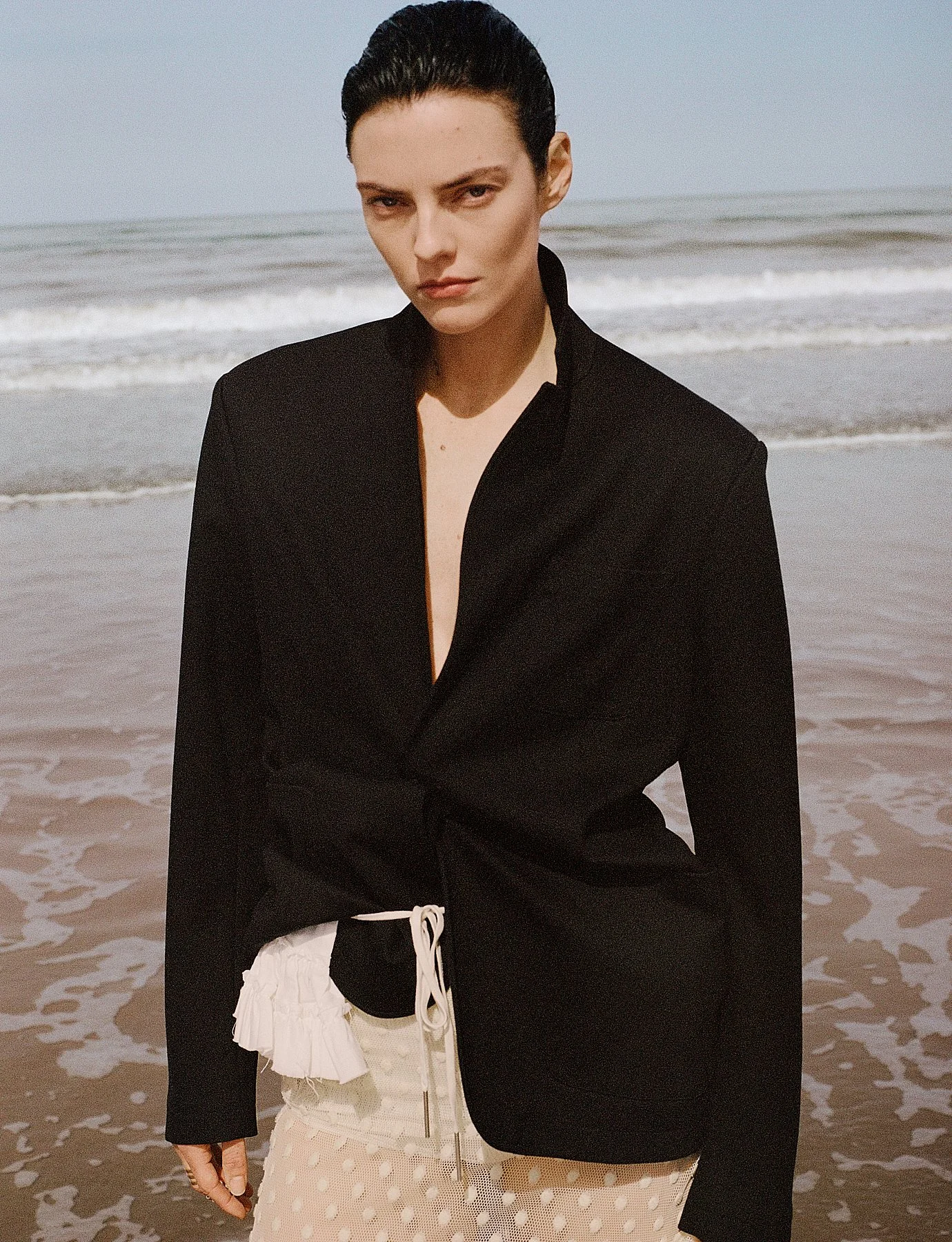 A woman with dark hair tied back, wearing a black blazer over a sheer polka dot dress, stands on a beach with the ocean in the background.