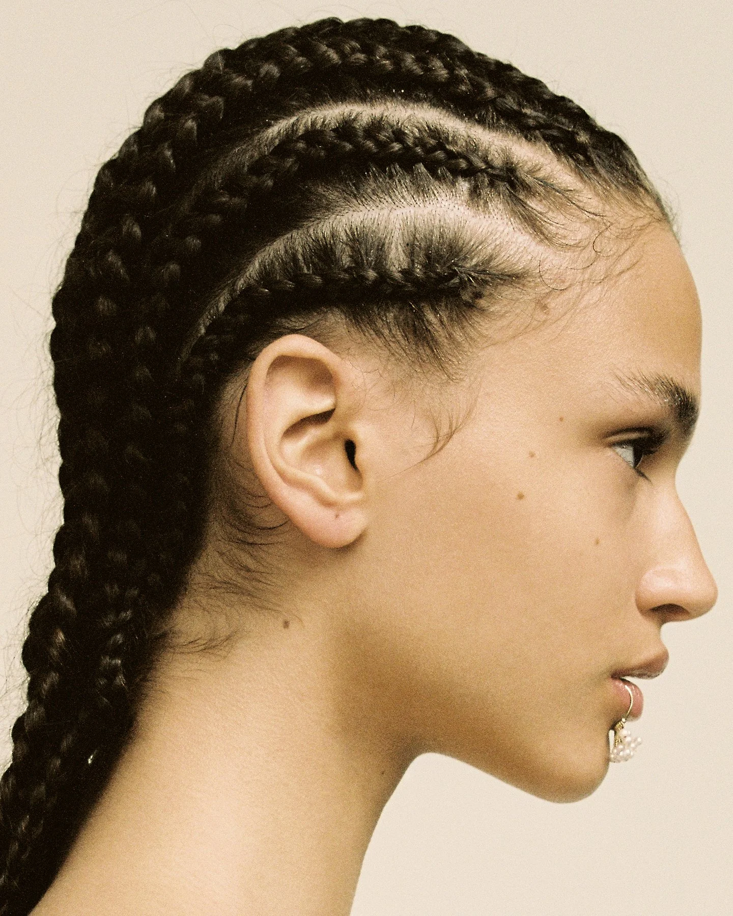 A woman with braided hair, earrings, and a nose ring in profile against a plain background.