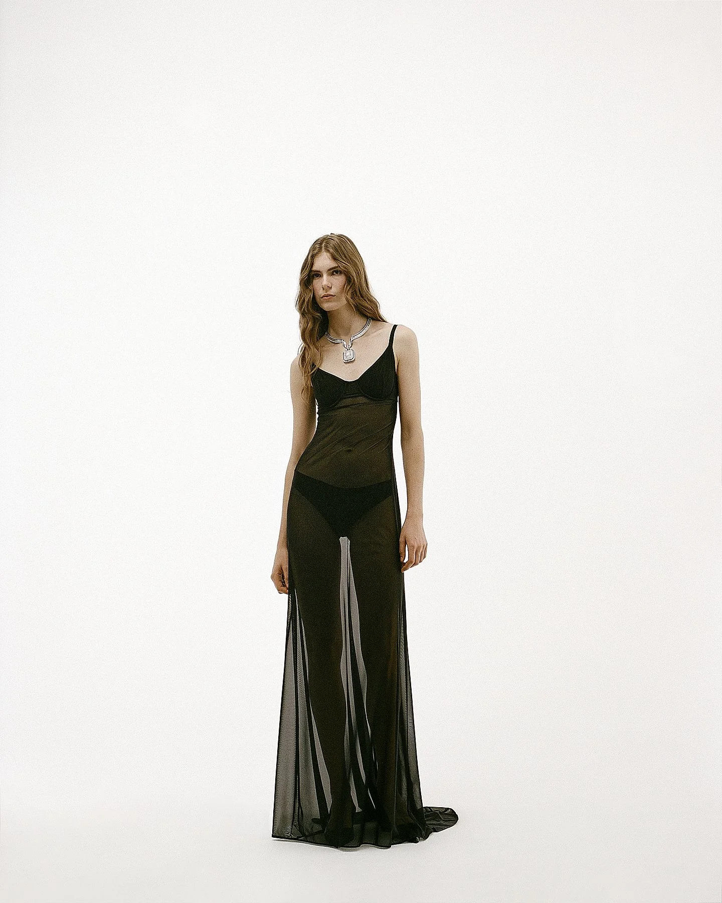 A young woman with long, wavy brown hair wearing a black sheer dress with thin straps and a slit, paired with a large silver necklace, standing against a plain white background.