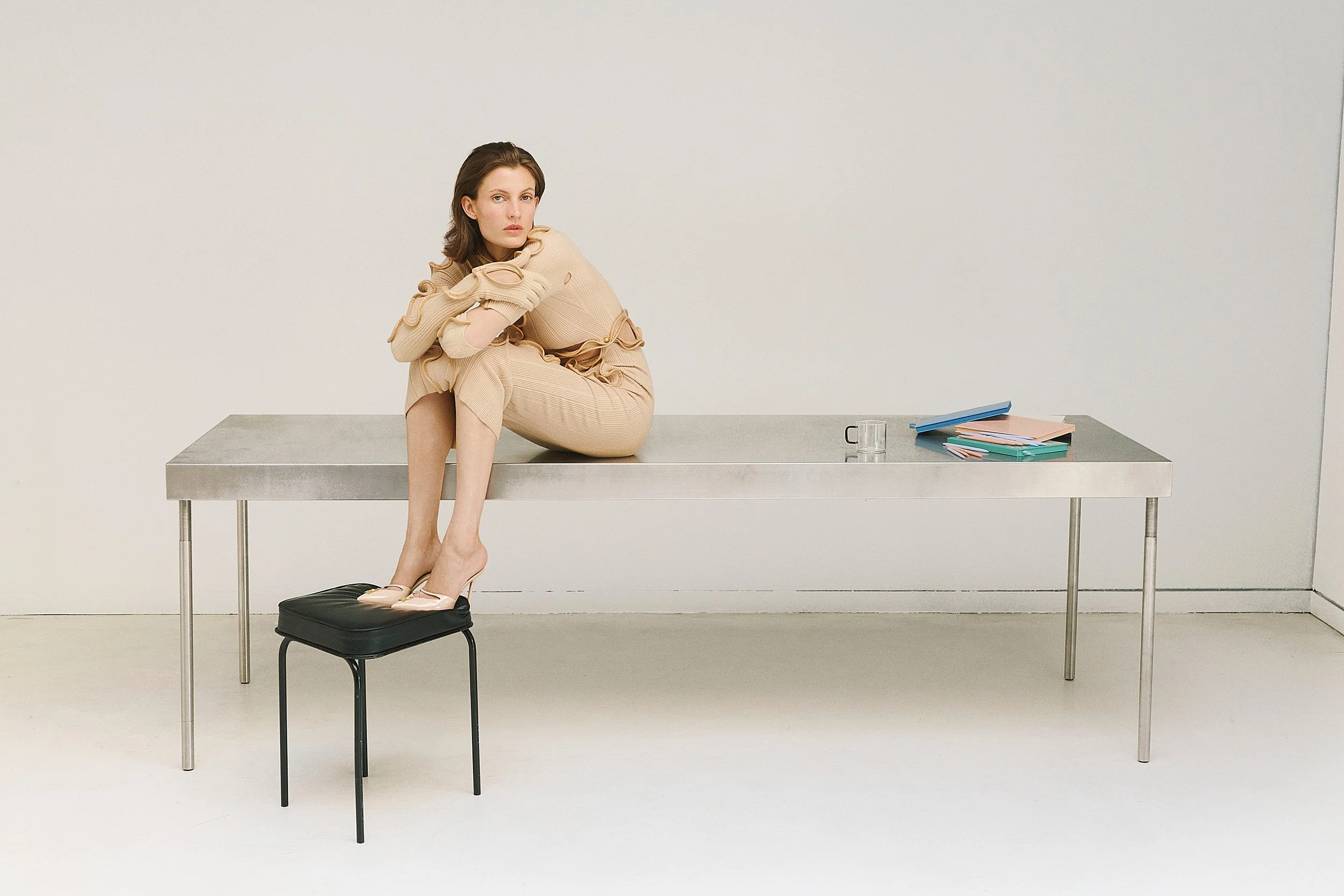 A woman sitting on a metallic table in a minimalistic room with a plain white wall. She wears beige clothing with cut-out details, has brown hair, and is barefoot with her shoes on a black stool in front of her. There is a stack of colorful notebooks