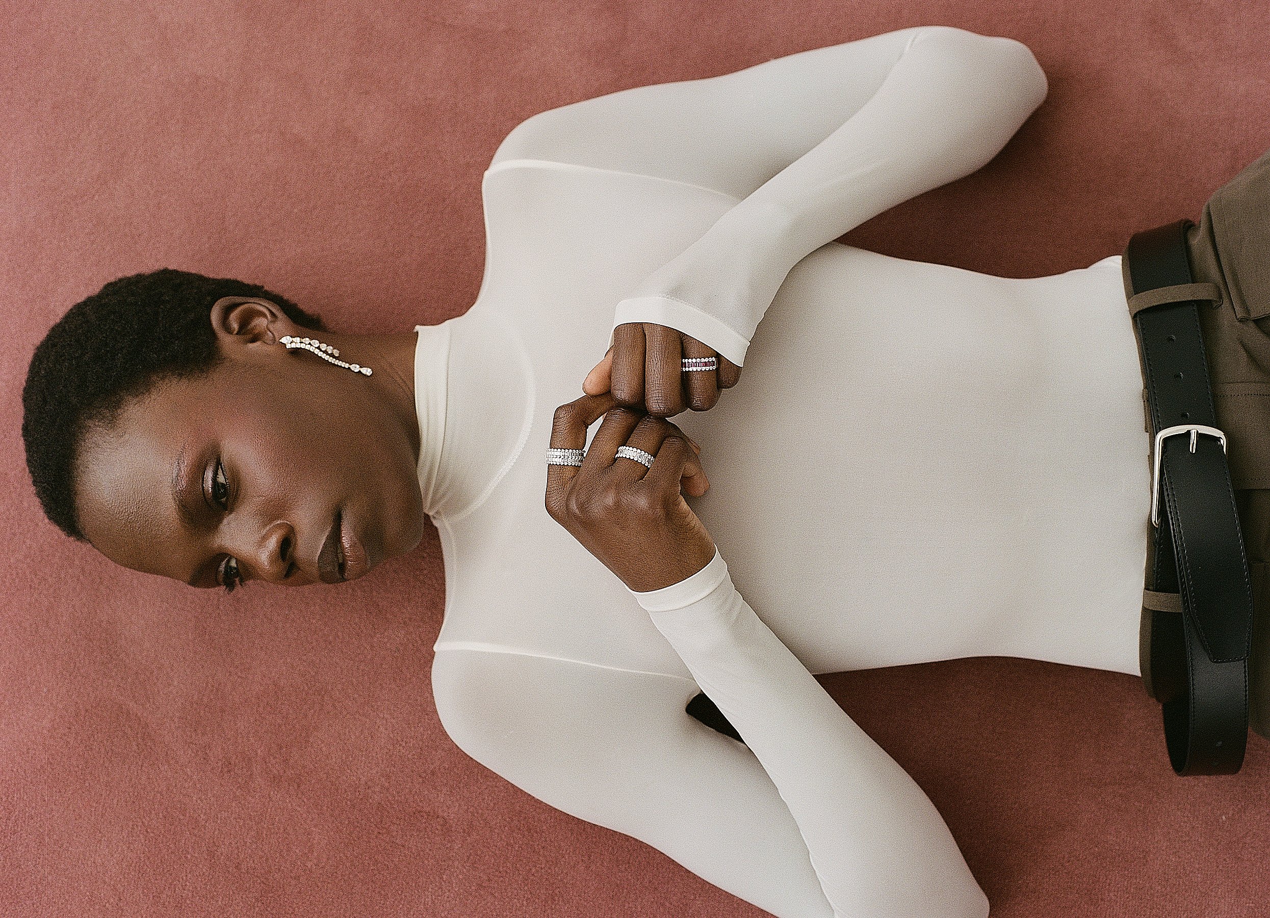 A woman with short curly hair lying on the floor with her eyes closed, wearing a white long-sleeve top, diamond earrings, and rings, with a black belt and beige pants, against a brown background.